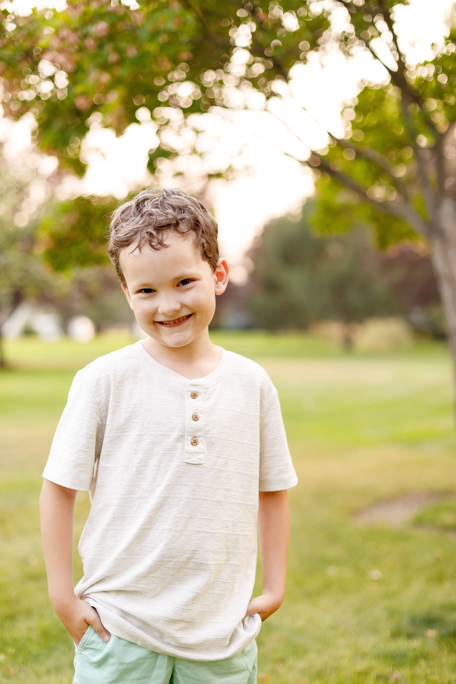 A child wearing casual clothes smiles warmly while standing in a park setting.