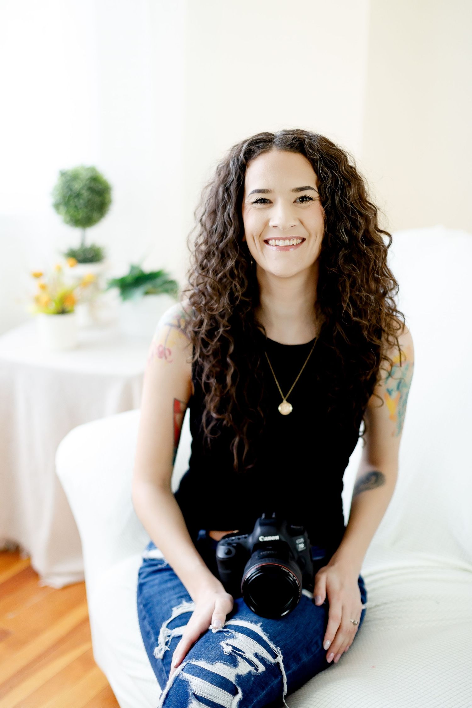 Professional portrait series showing a relaxed person in casual black top and jeans sitting on white furniture in a bright room.