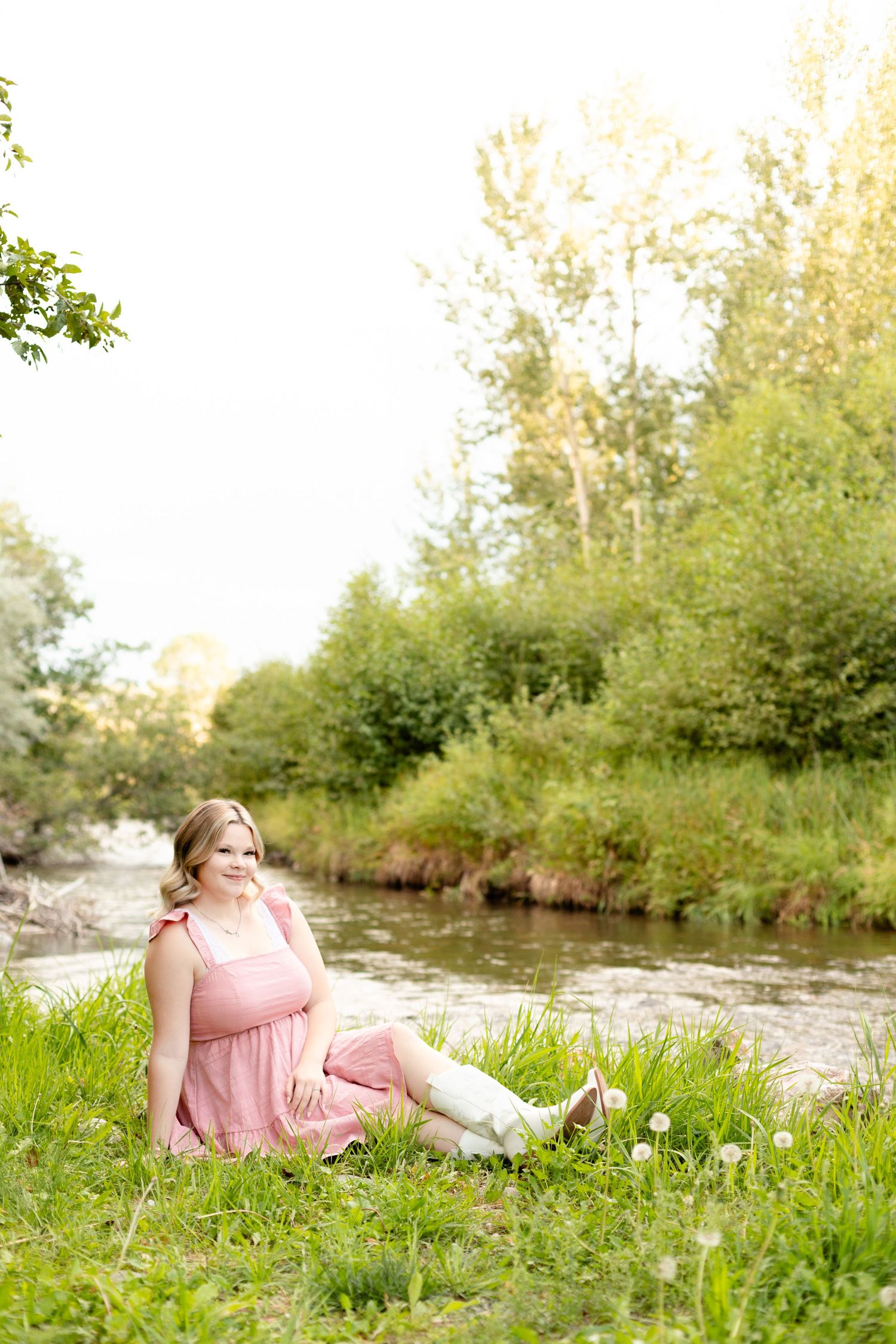 A person in a pink dress relaxes by a stream on a grassy bank during golden hour.