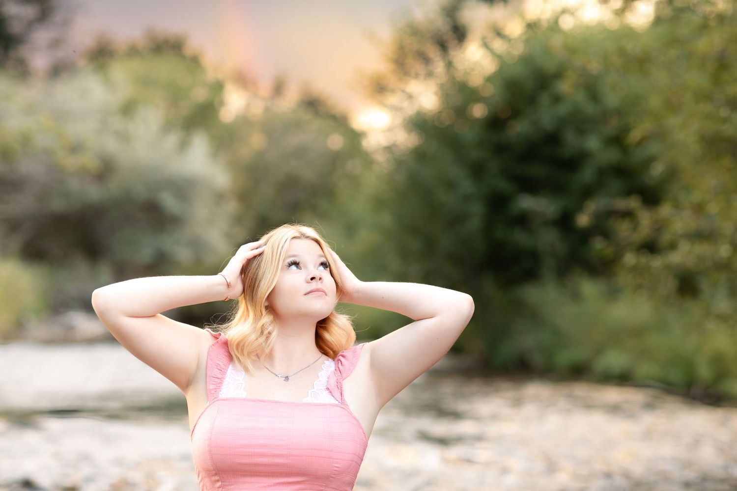 A woman in a pink top poses outdoors during golden hour with soft natural lighting and blurred foliage background.