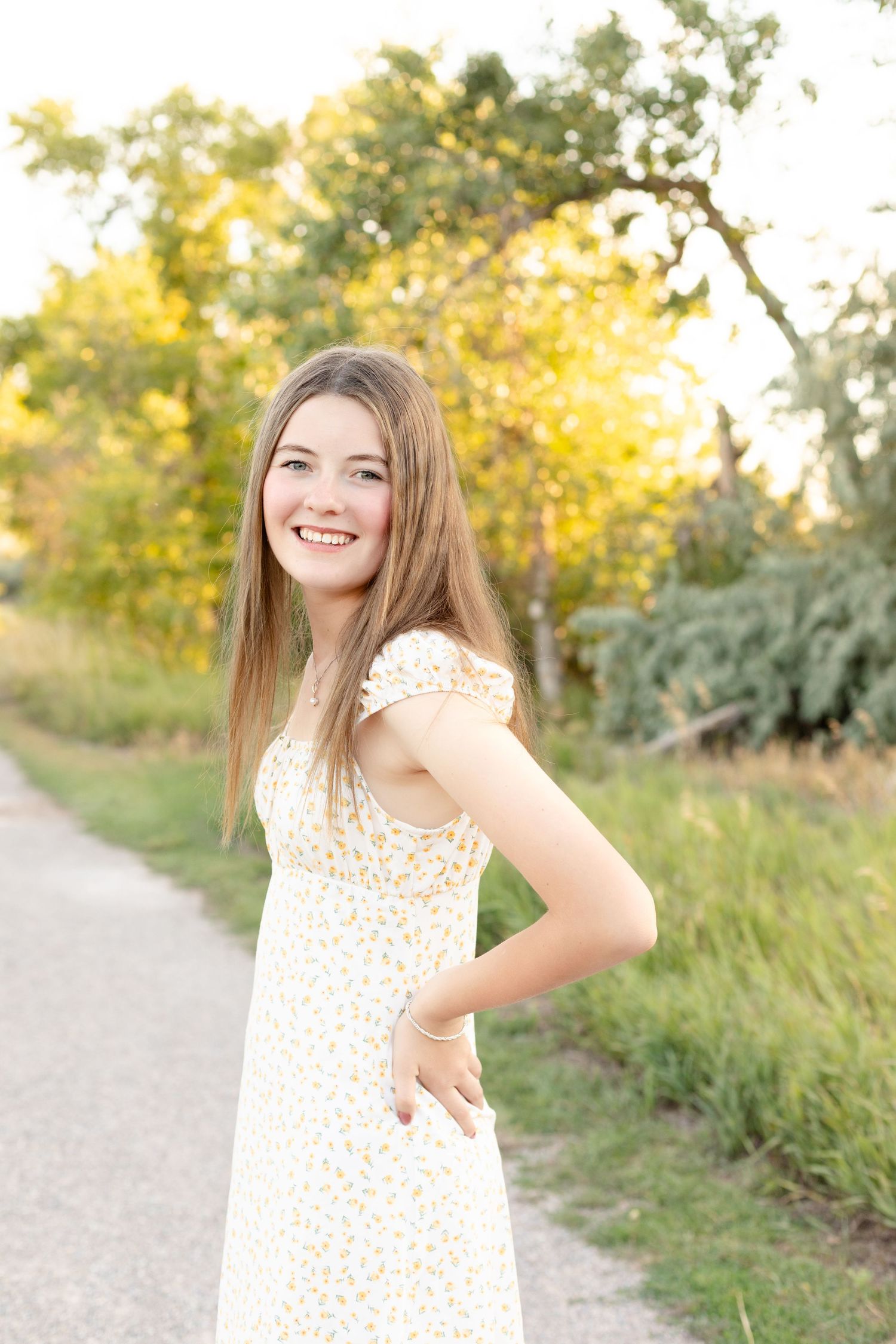 Person wearing a floral dress poses outdoors against a backdrop of autumn trees and natural scenery.