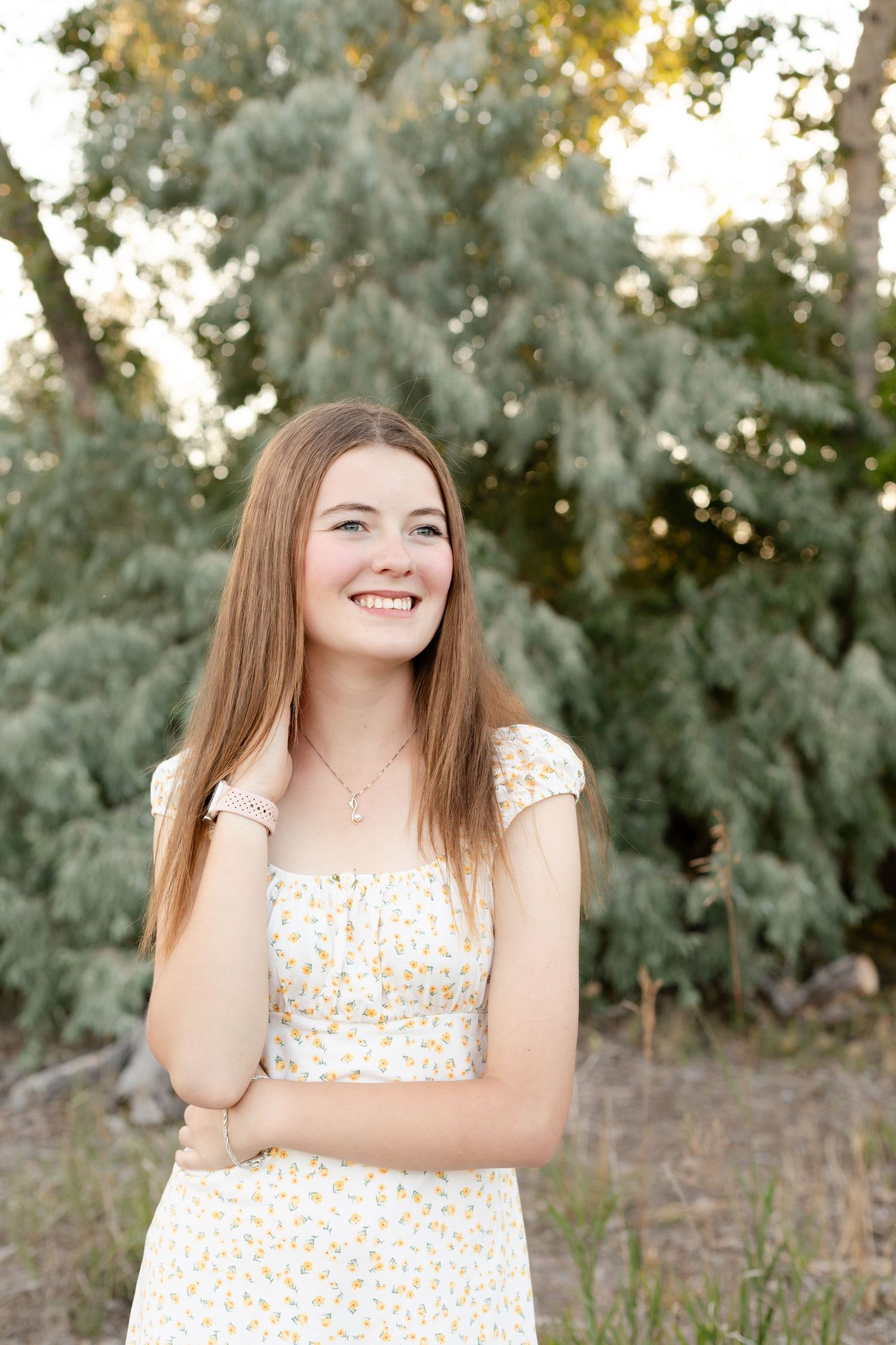 A person in a white floral dress smiles joyfully while posing outdoors against a natural green background.