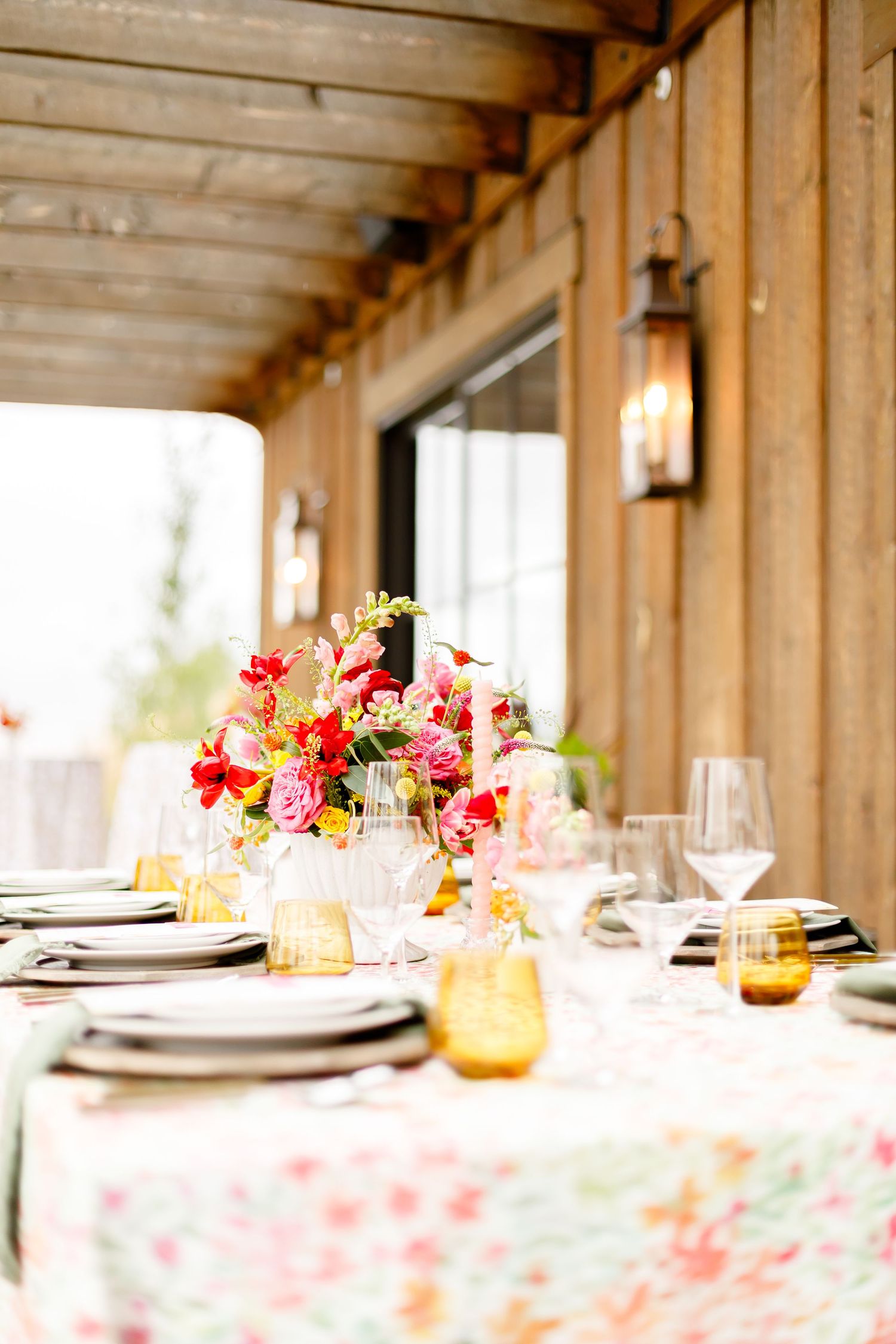 Elegant table setting with floral tablecloth, amber glassware and vibrant pink flower centerpiece on rustic wooden porch.