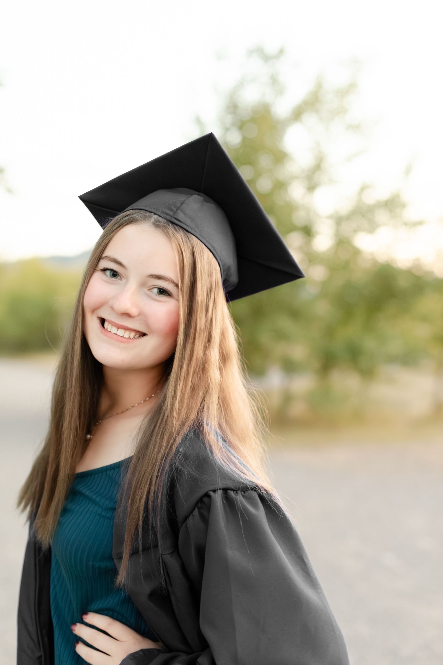 Graduate in dark blue sweater and black cap poses outdoors against blurred natural backdrop.