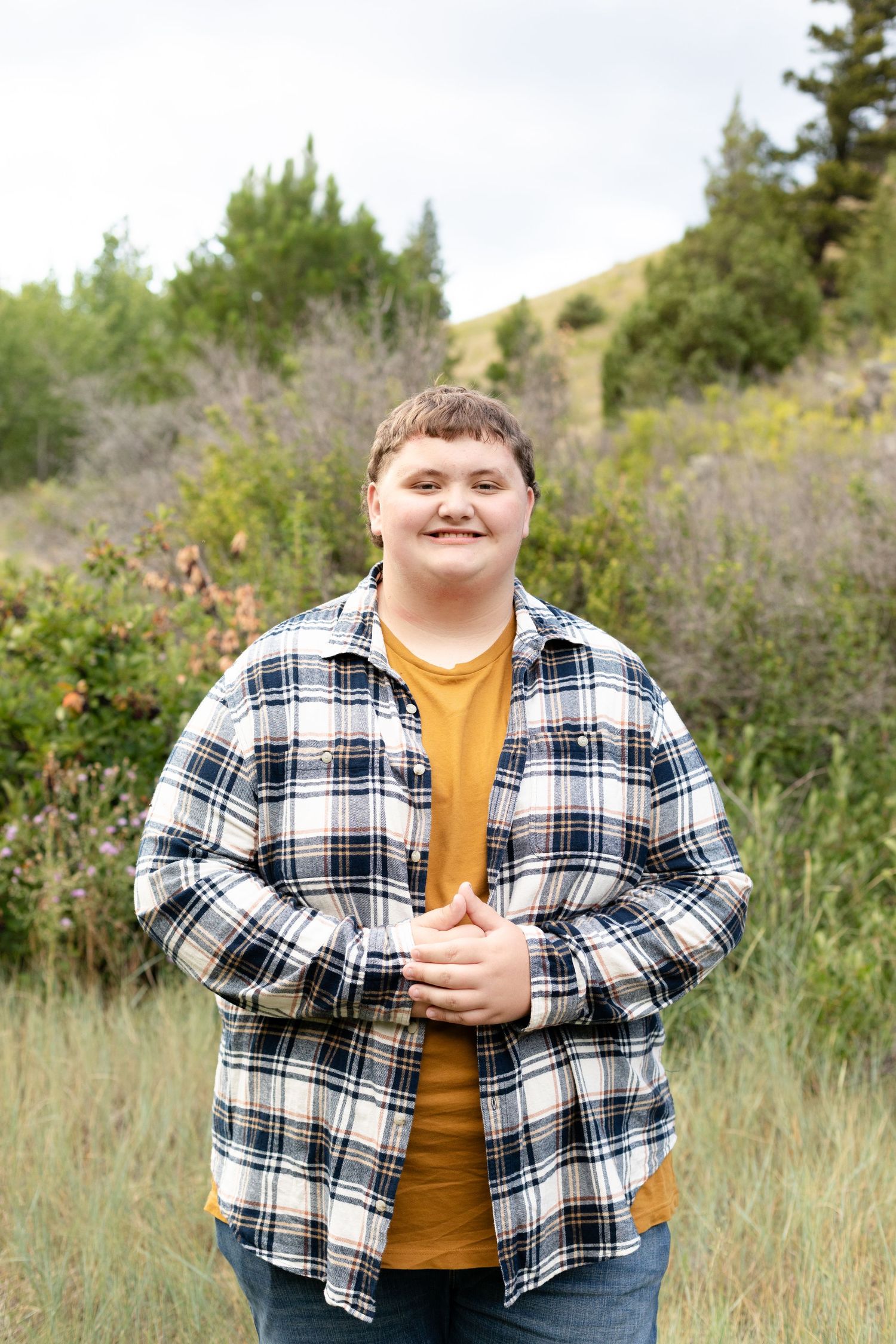 A person wearing a plaid flannel shirt and yellow t-shirt poses in grassy outdoor setting with natural foliage.