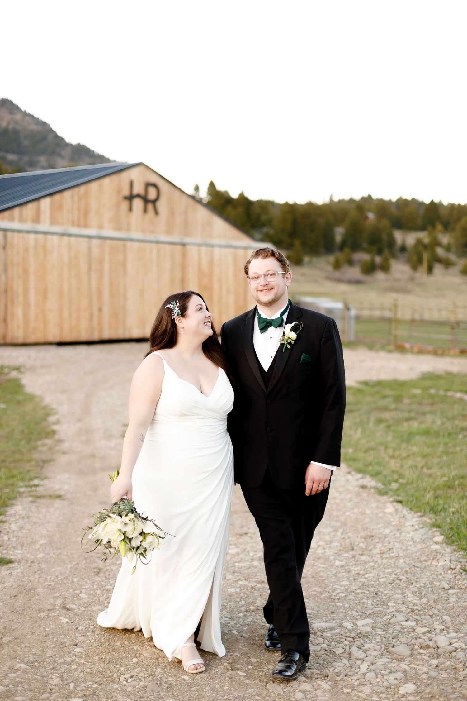 A bride and groom walk together on a dirt path in front of a rustic barn with mountains in the background.