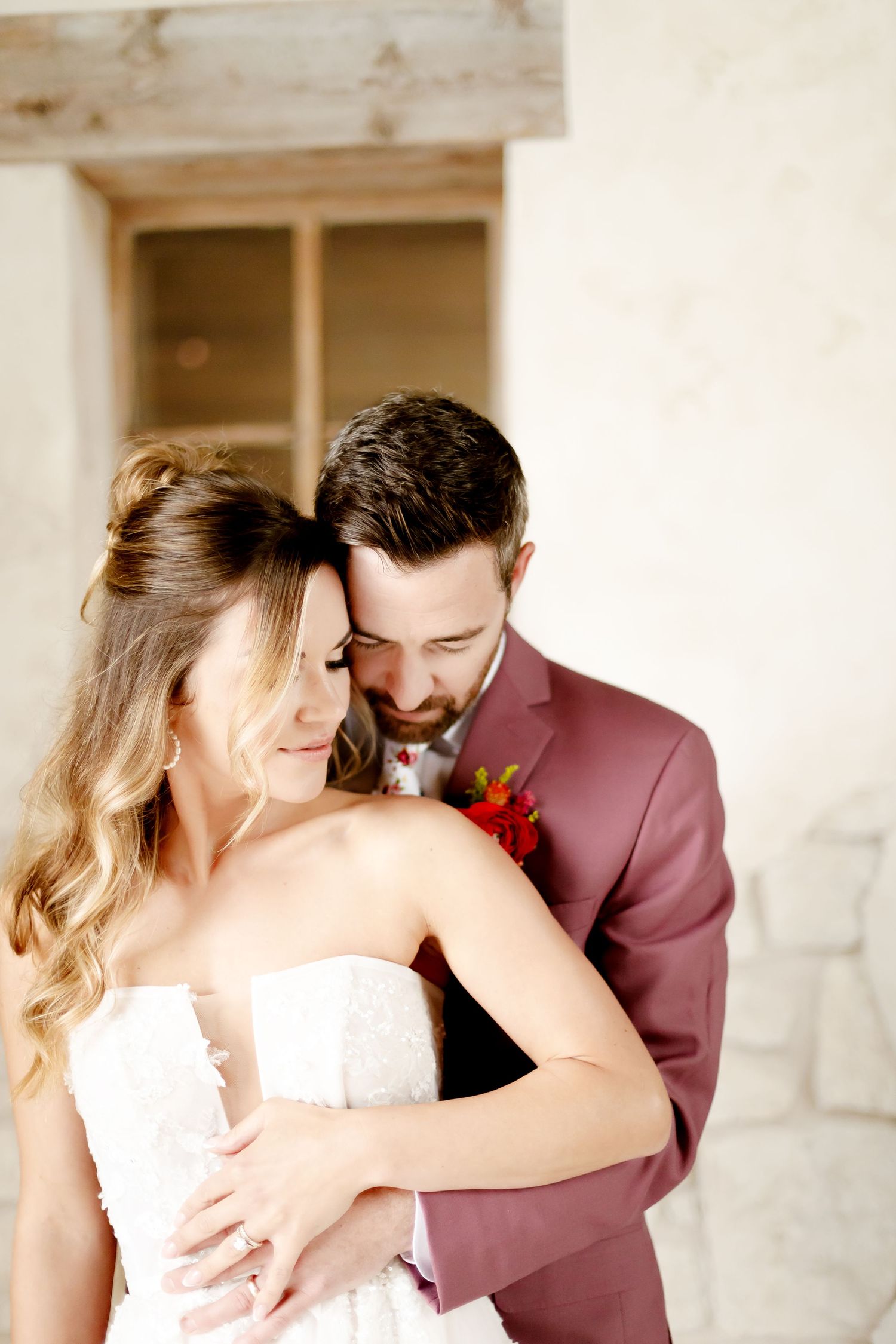 Romantic wedding couple embracing in an intimate moment against a light colored wall, wearing mauve suit and white dress.