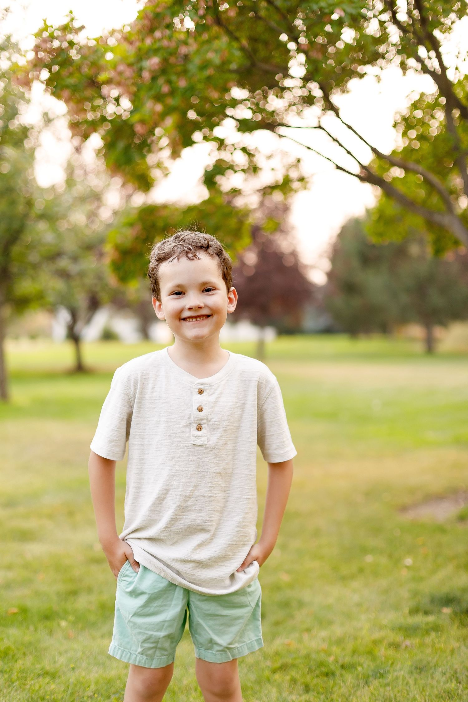 A child in a beige henley shirt and mint shorts stands in a lush green park.