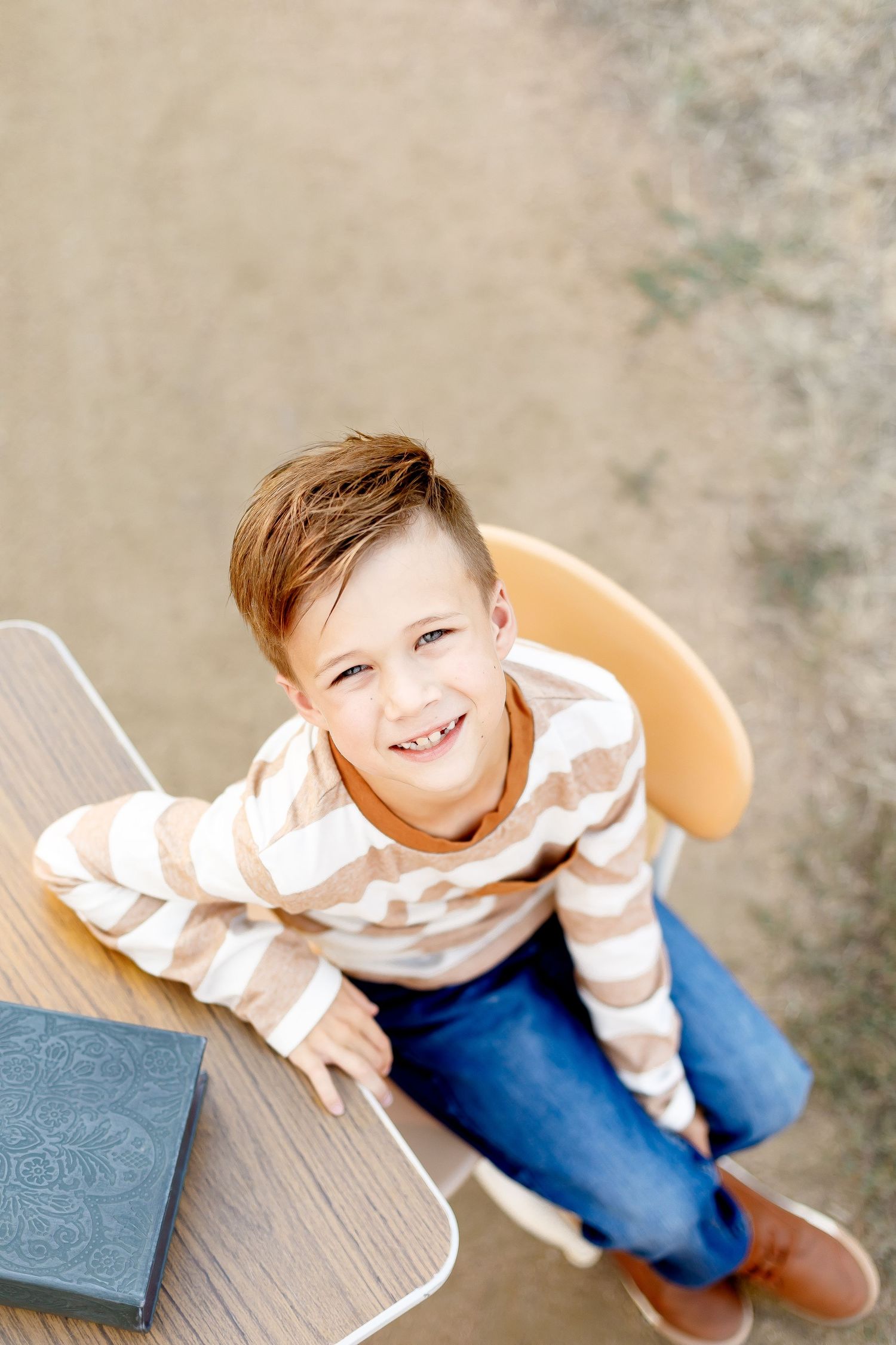 A child in a striped sweater and jeans sits on wooden decking while looking up at the camera.
