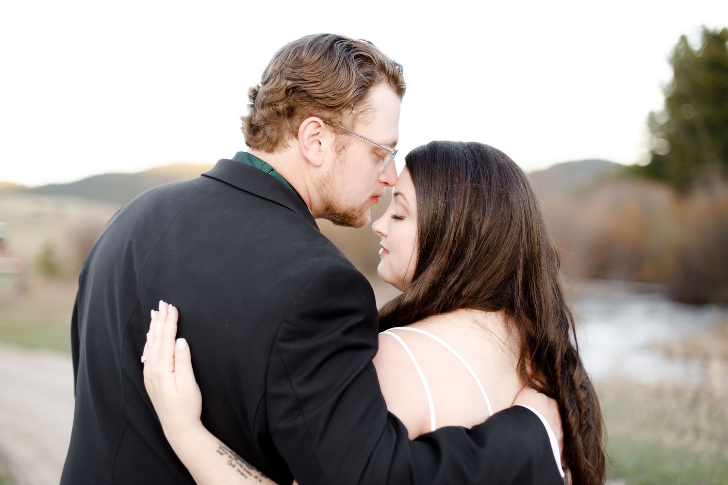 A couple shares a romantic moment outdoors at sunset as they embrace in elegant formal attire.