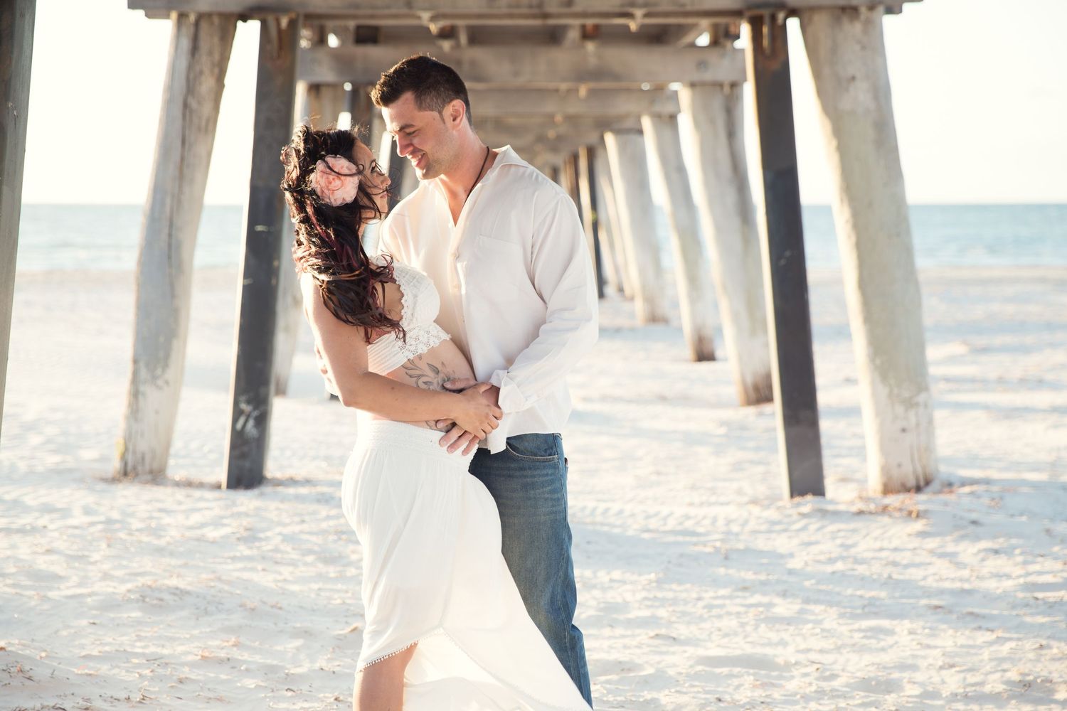A lady in a crochet bra and flowy skirt in a romantic embrace under the Largs Bay jetty for a maternity photoshoot.