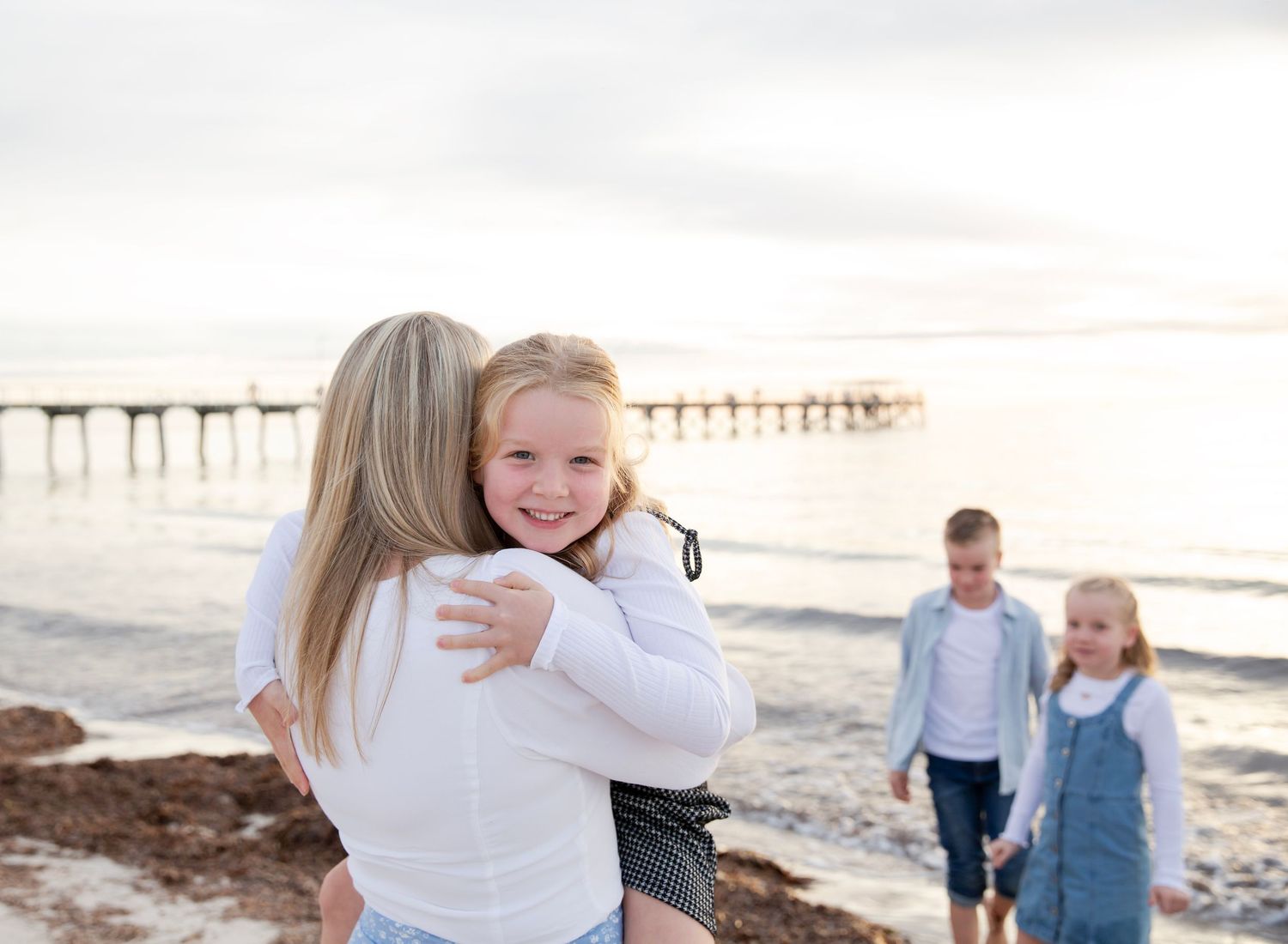 daughter cuddles mum and looks cheekily at the camera while her siblings play in the background