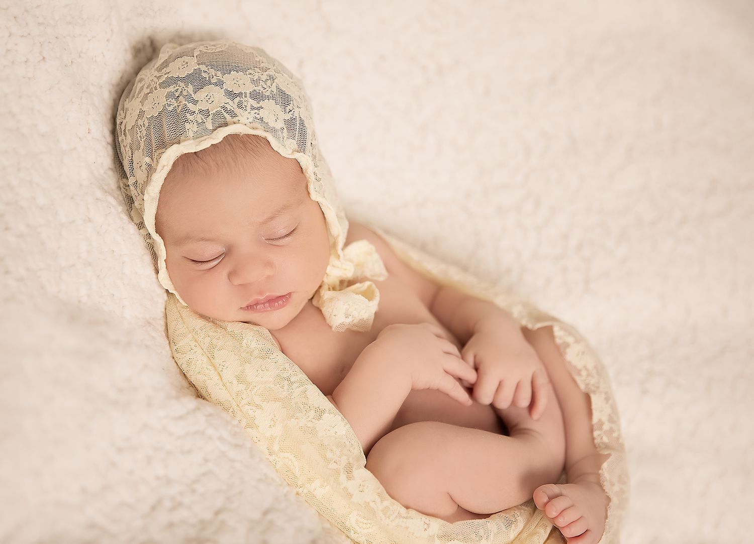 A newborn baby girl in a lace wrap and bonnet, sleeping soundly with her feet tucked up in in home newborn photography session.