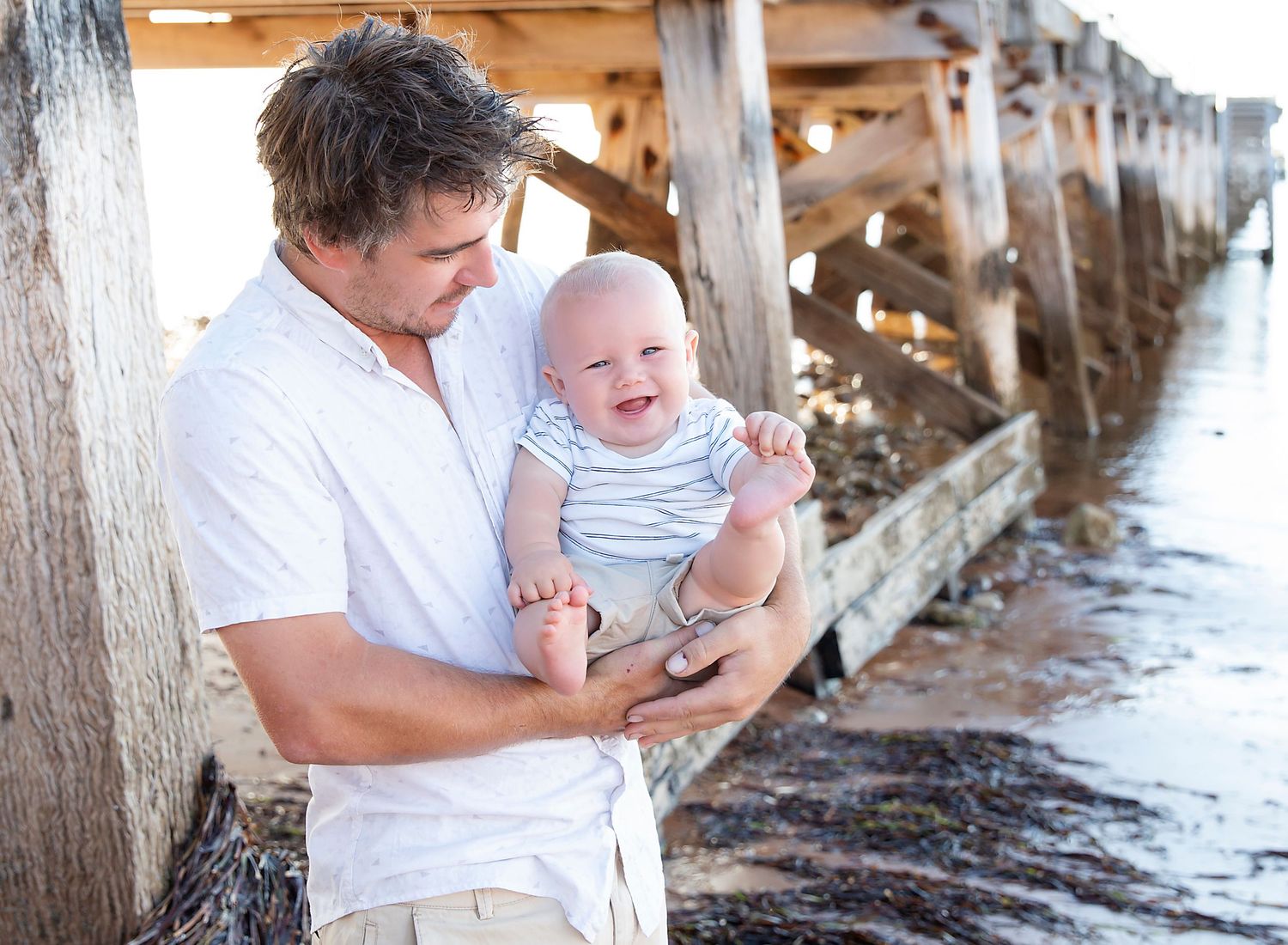A dad and his 6 month old baby who is smiling in family photoshoot at the beach jetty in Ardrossan SA.