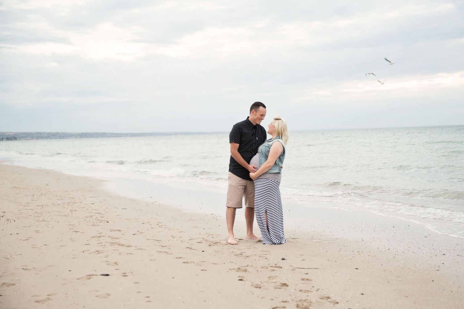 A couple hugging their baby bump and smiling at each other in their maternity photoshoot at the beach in Henley Beach South SA.