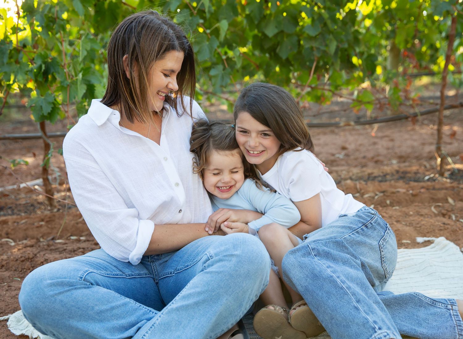 A mum laughs as her two daughters cuddle each other lovingly in family photo session in vineyard in Angle Vale SA.
