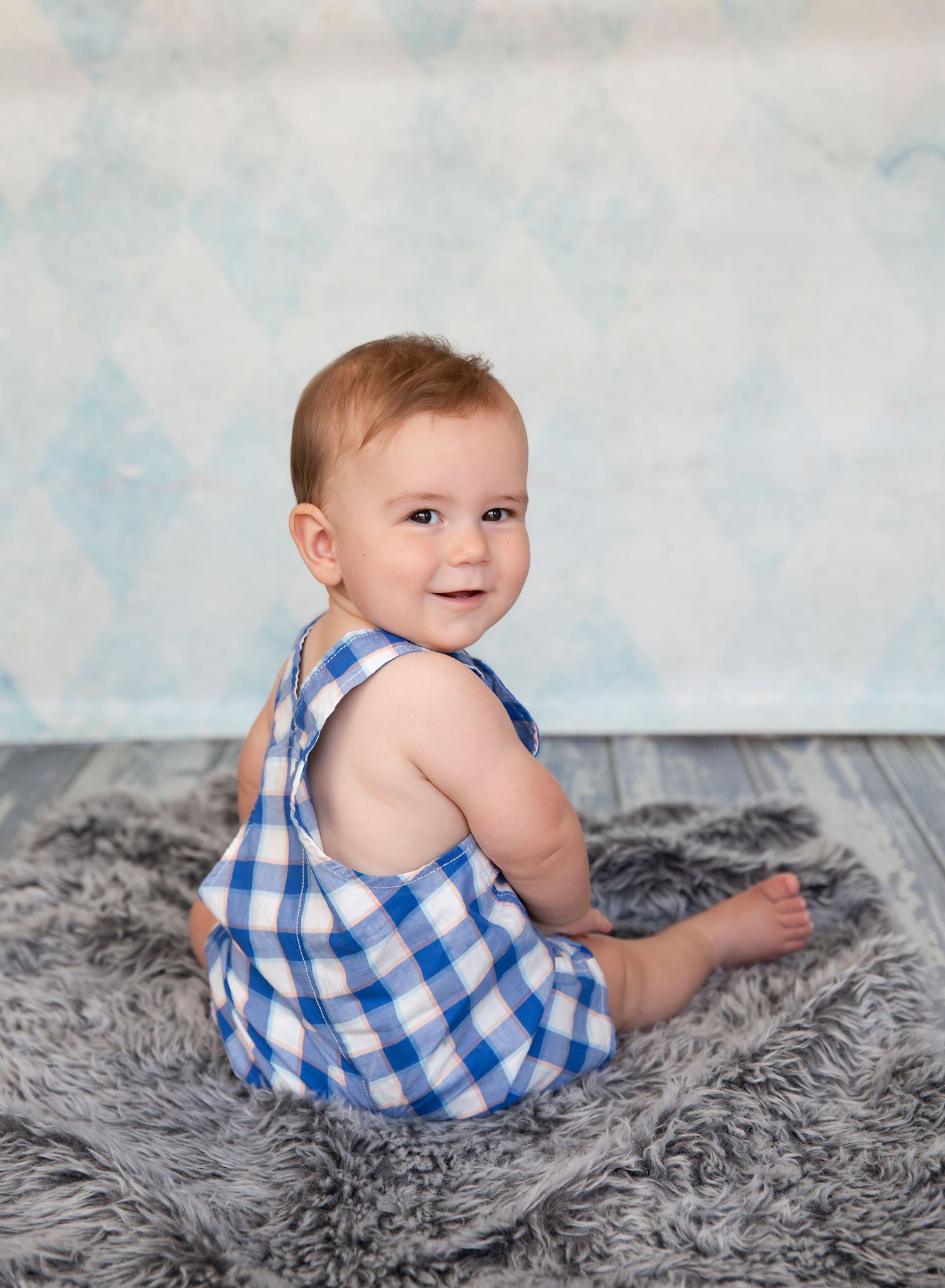 7 month old baby in overalls sitting on a rug and smiling at the camera for his sitter session. In-home studio photoshoot in Adelaide SA.