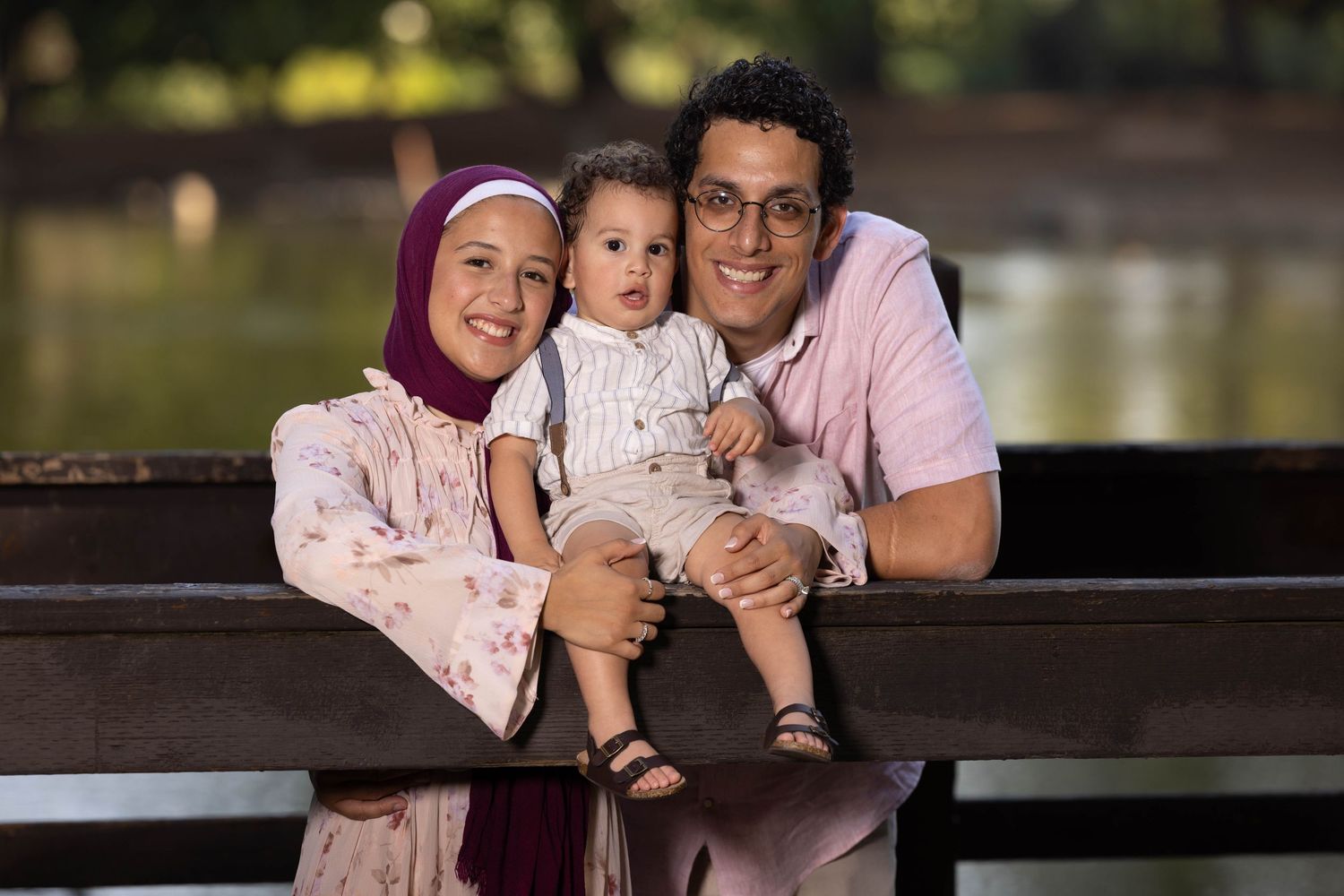 Smiling family portrait by the water in Fresno, captured by FresLens, featuring parents holding their toddler on a wooden bridge