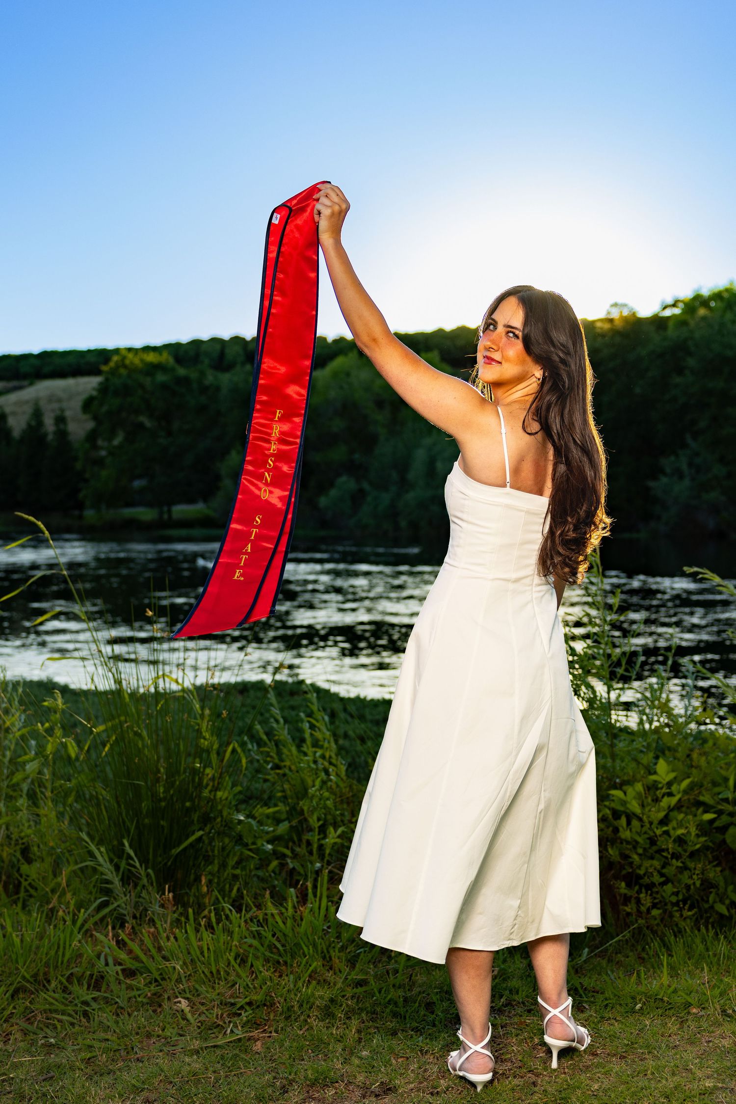Fresno state graduate, posing with red stole and white dress at Lost Lake in Fresno California.