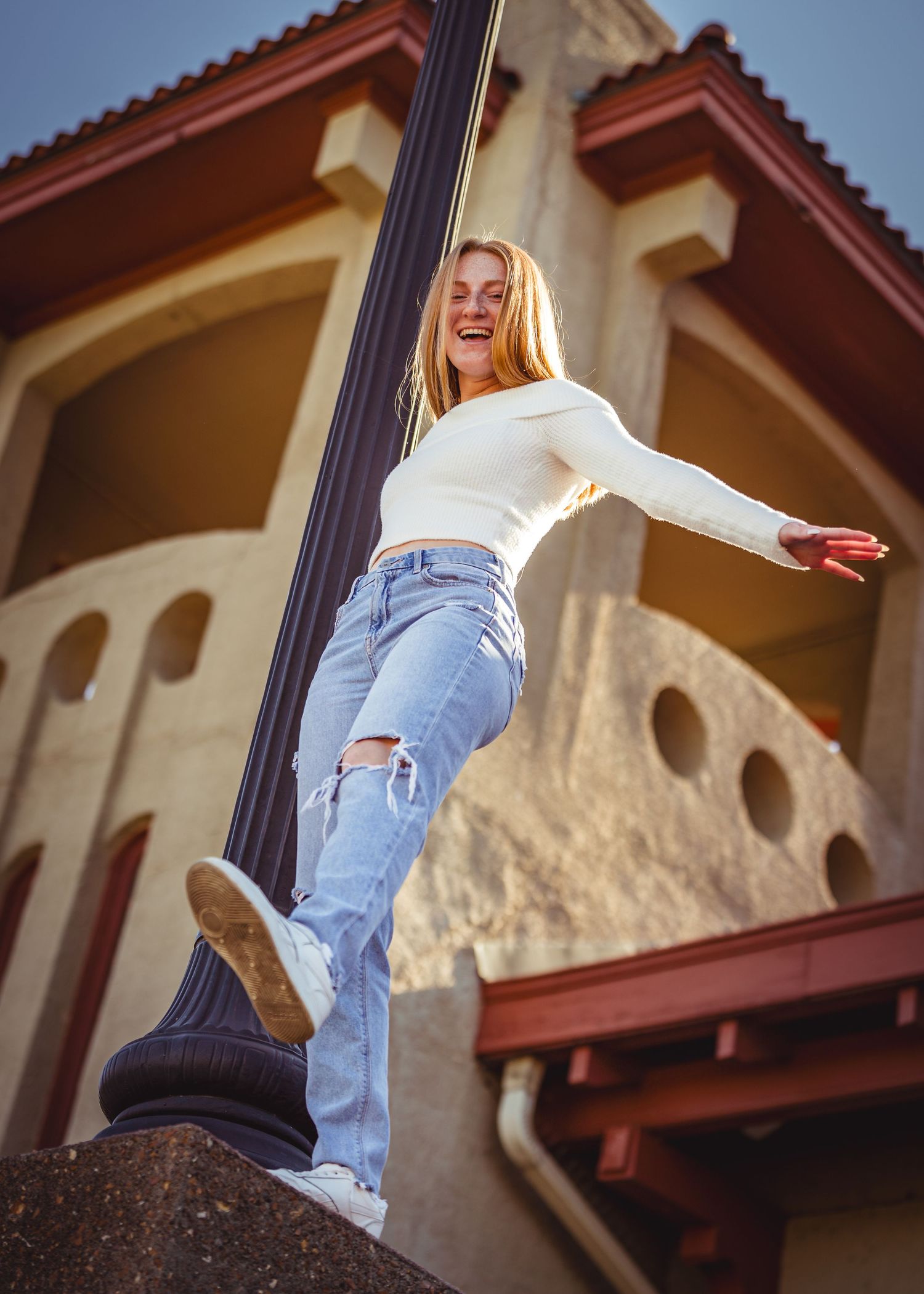 Person in ripped jeans and white top poses playfully against Mediterranean-style architecture.
