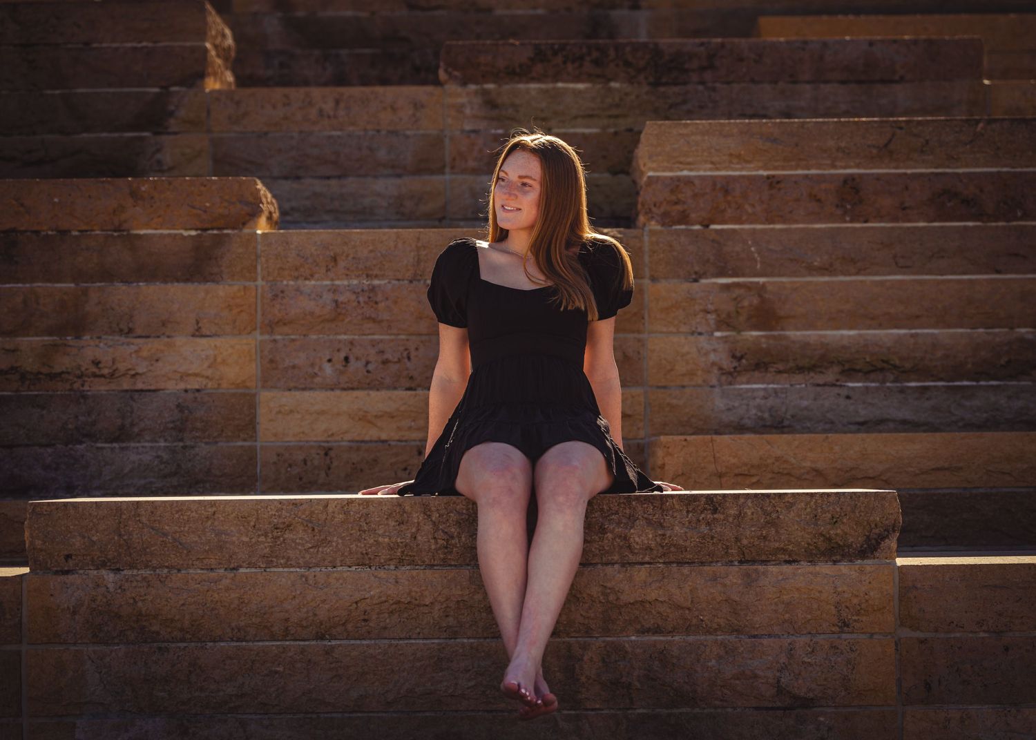 A person in a black dress sits contemplatively on stone steps in warm lighting.