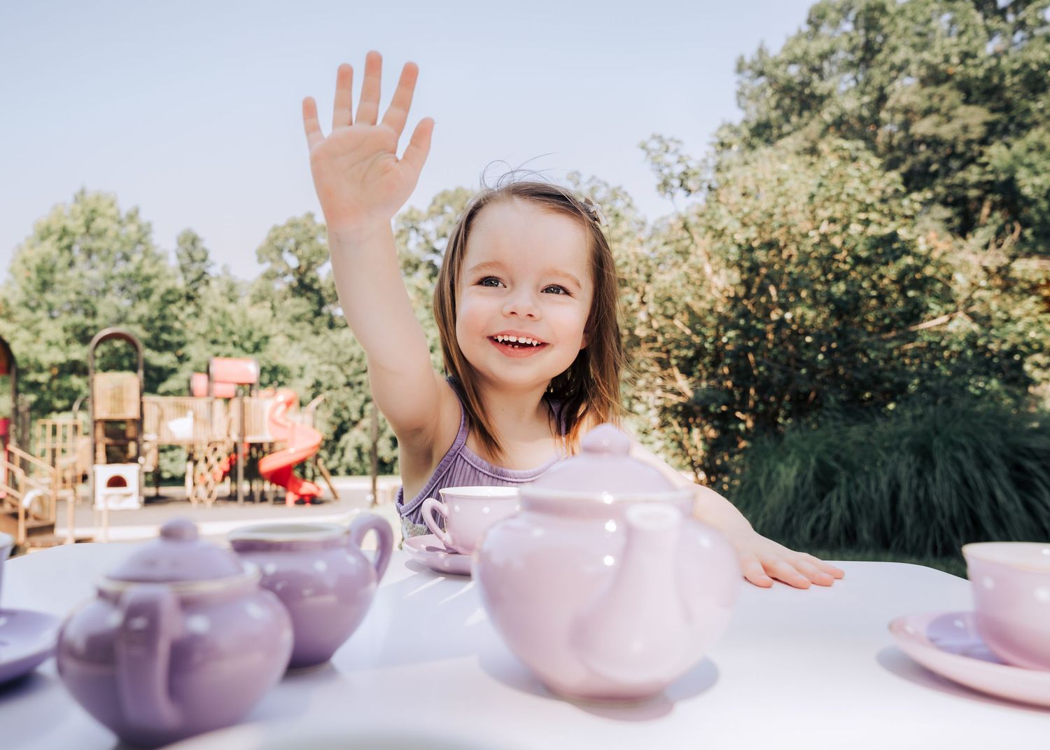A child waves while sitting at a pink tea party setup outdoors on a sunny day.