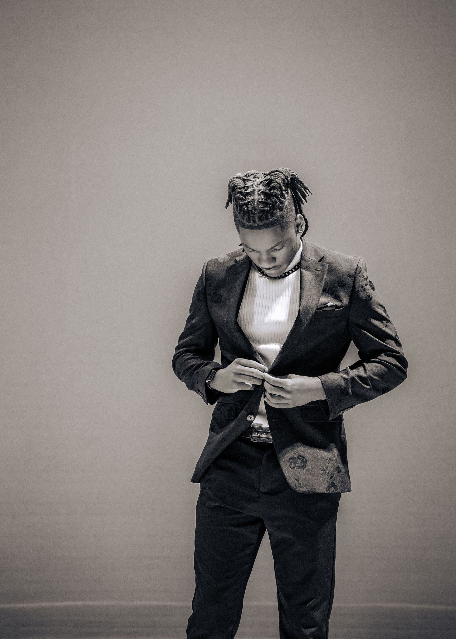 A fashionable figure in a black suit adjusts their clothing while looking downward in a monochromatic portrait.