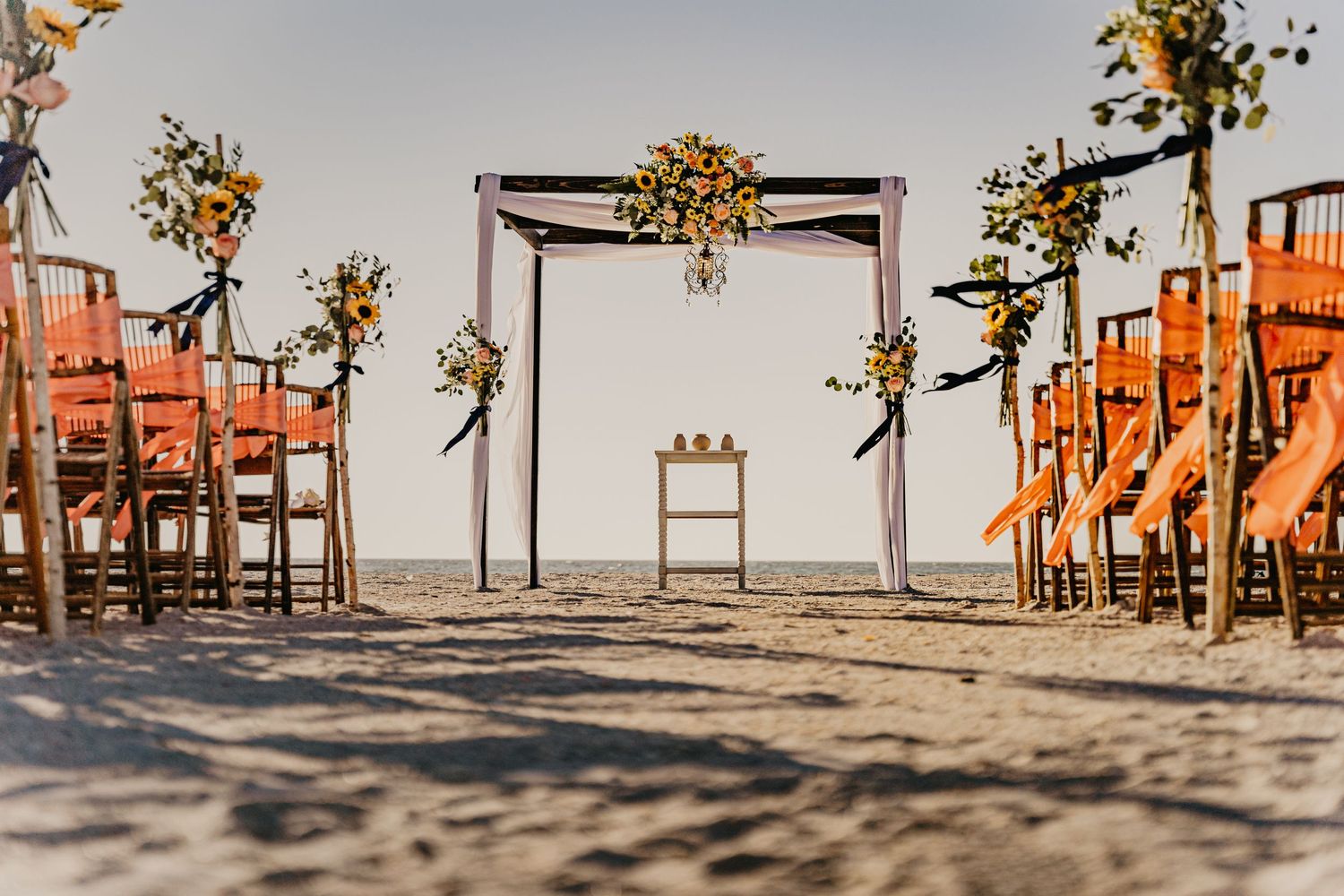 Beach wedding ceremony setup with wooden arch and orange chairs on sandy shore.