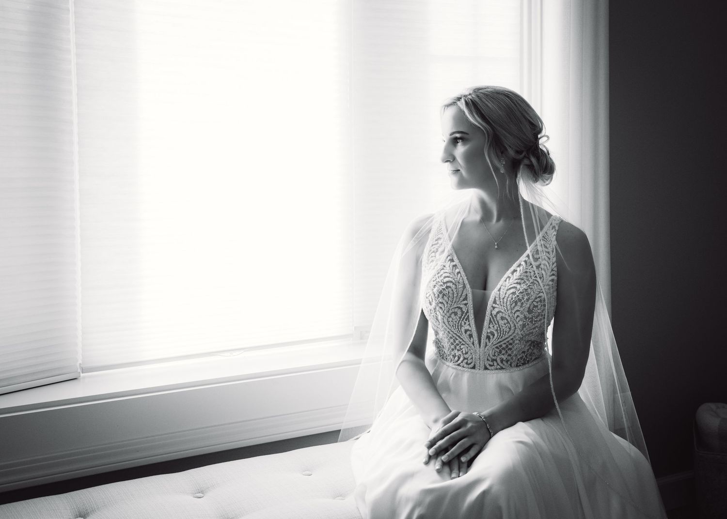 A bride in a deep v-neck gown poses elegantly by a window in black and white.
