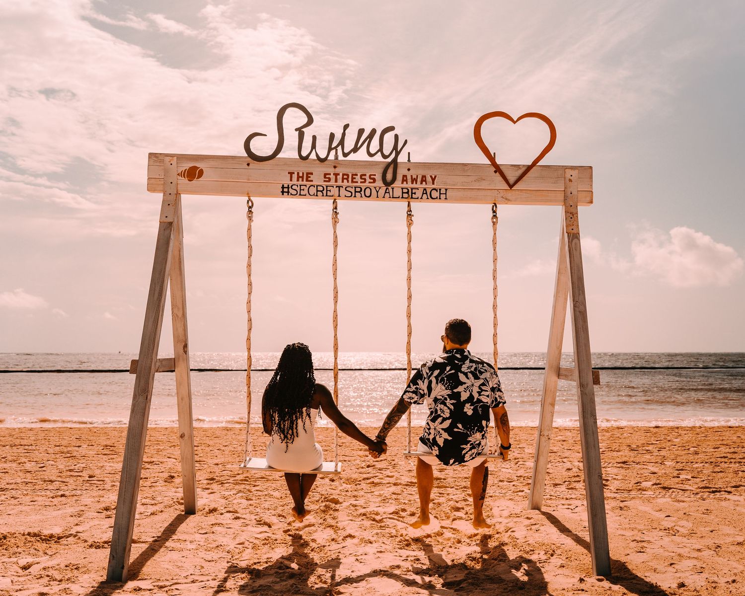 Silhouetted couple holding hands on a beach swing at sunset with heart decoration overhead.
