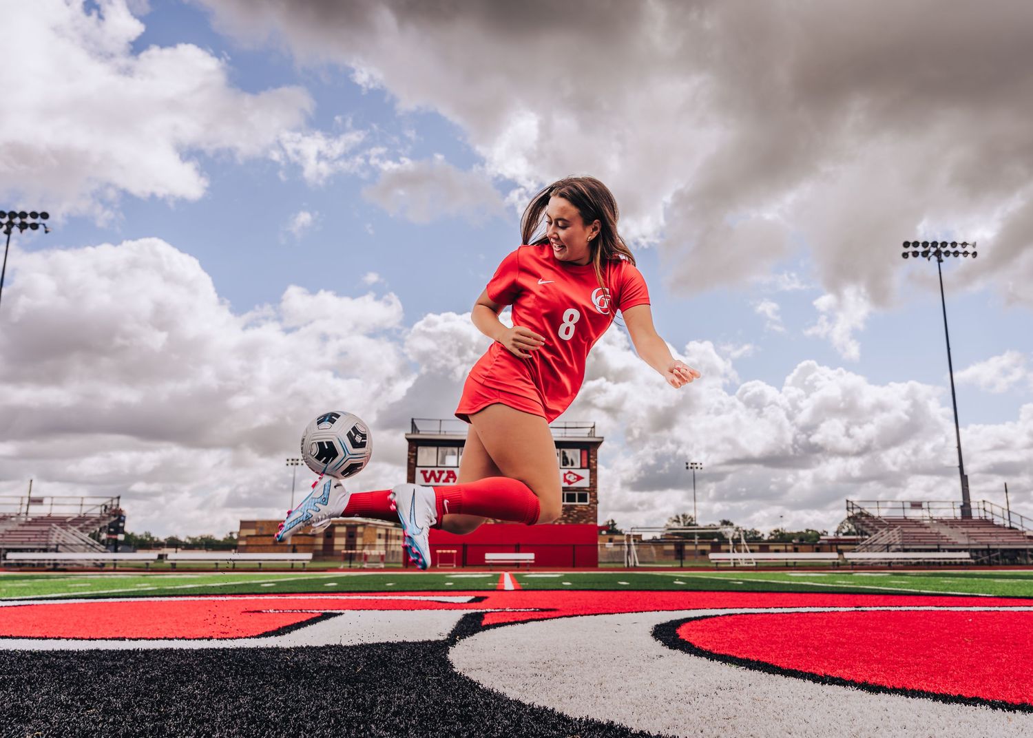 Soccer player in red uniform poses with ball on sports field under dramatic sky.