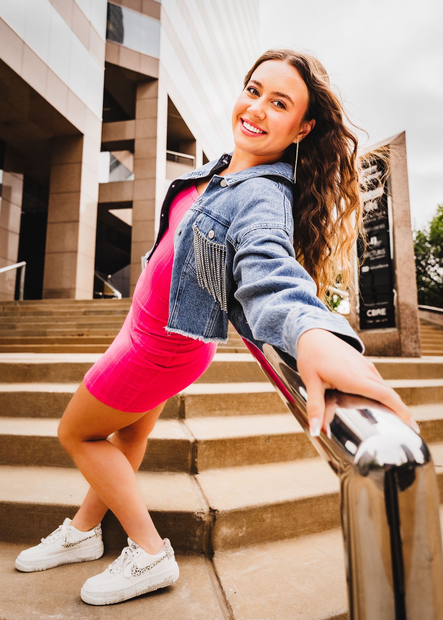 Student in pink dress and denim jacket poses playfully on campus steps.