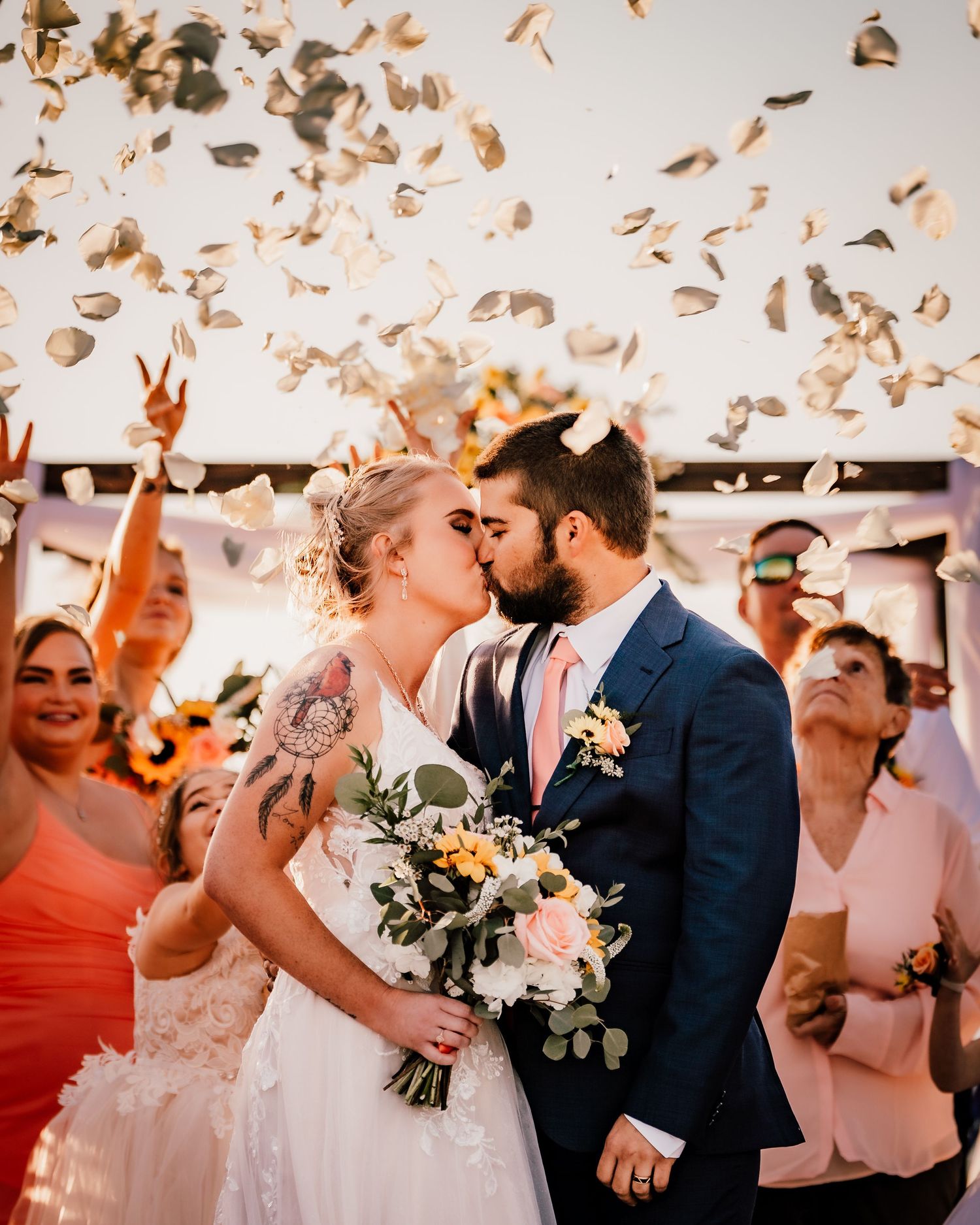 Wedding kiss captured during confetti celebration with coral-dressed guests.