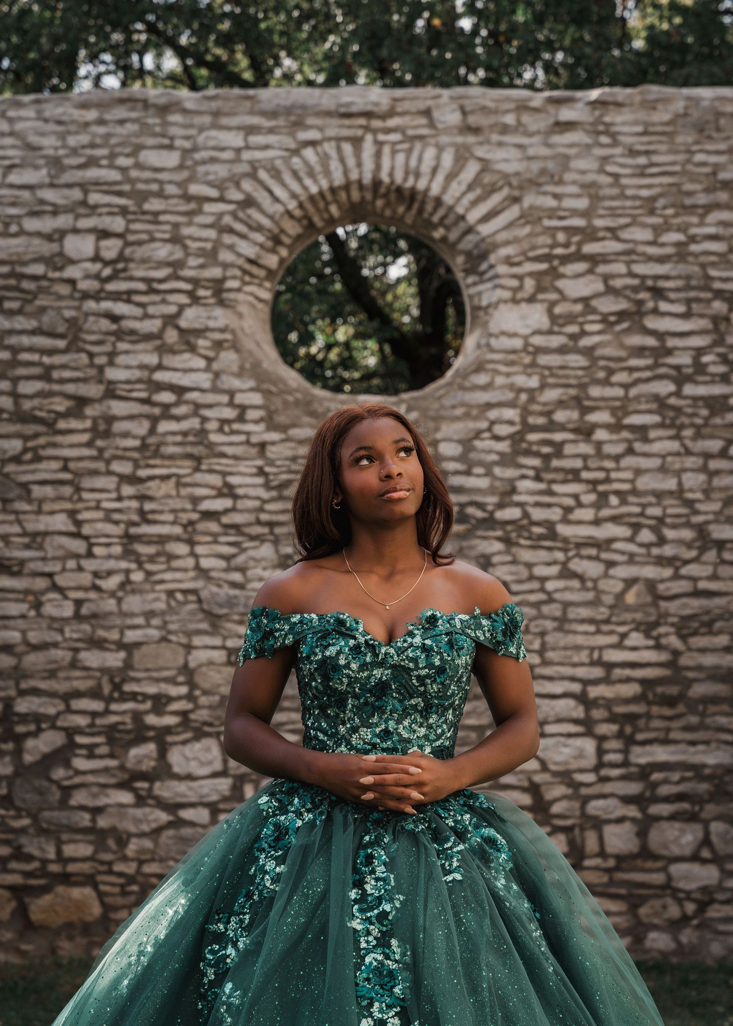 Elegant formal portrait against a stone wall with circular window wearing a sparkly teal gown.