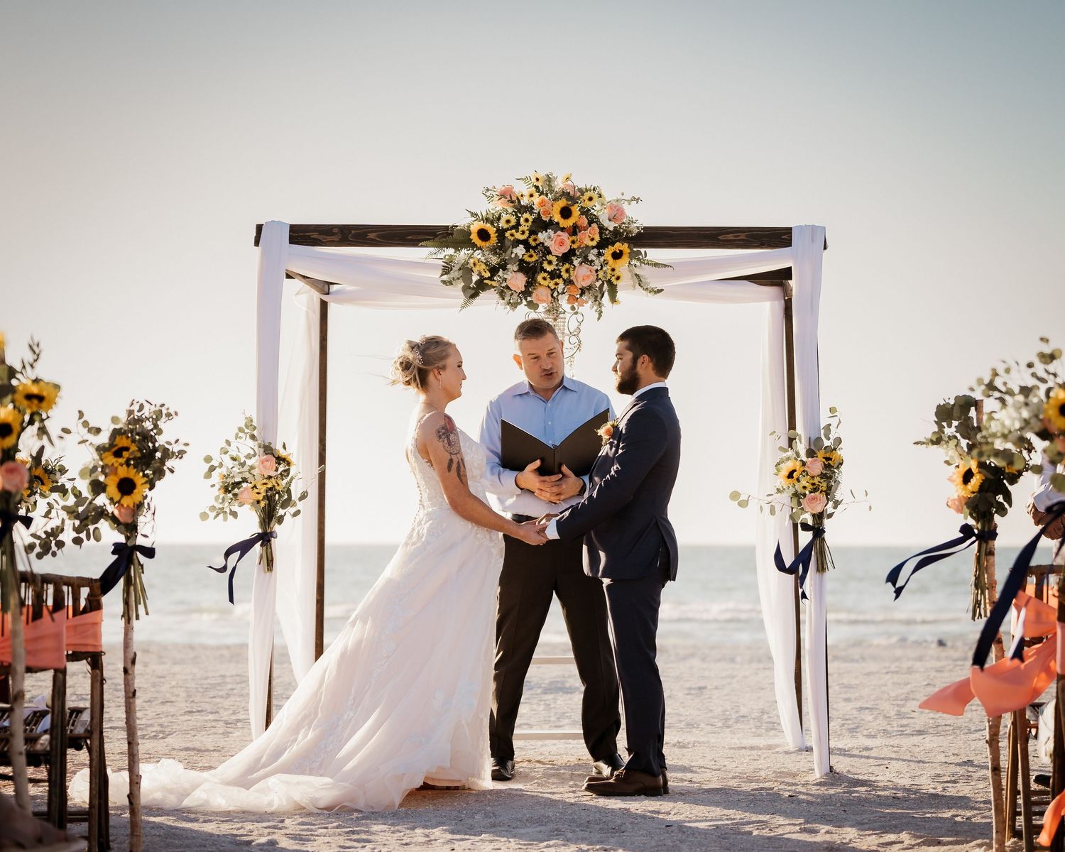 Beach wedding ceremony under a white arch decorated with floral arrangements at sunset.