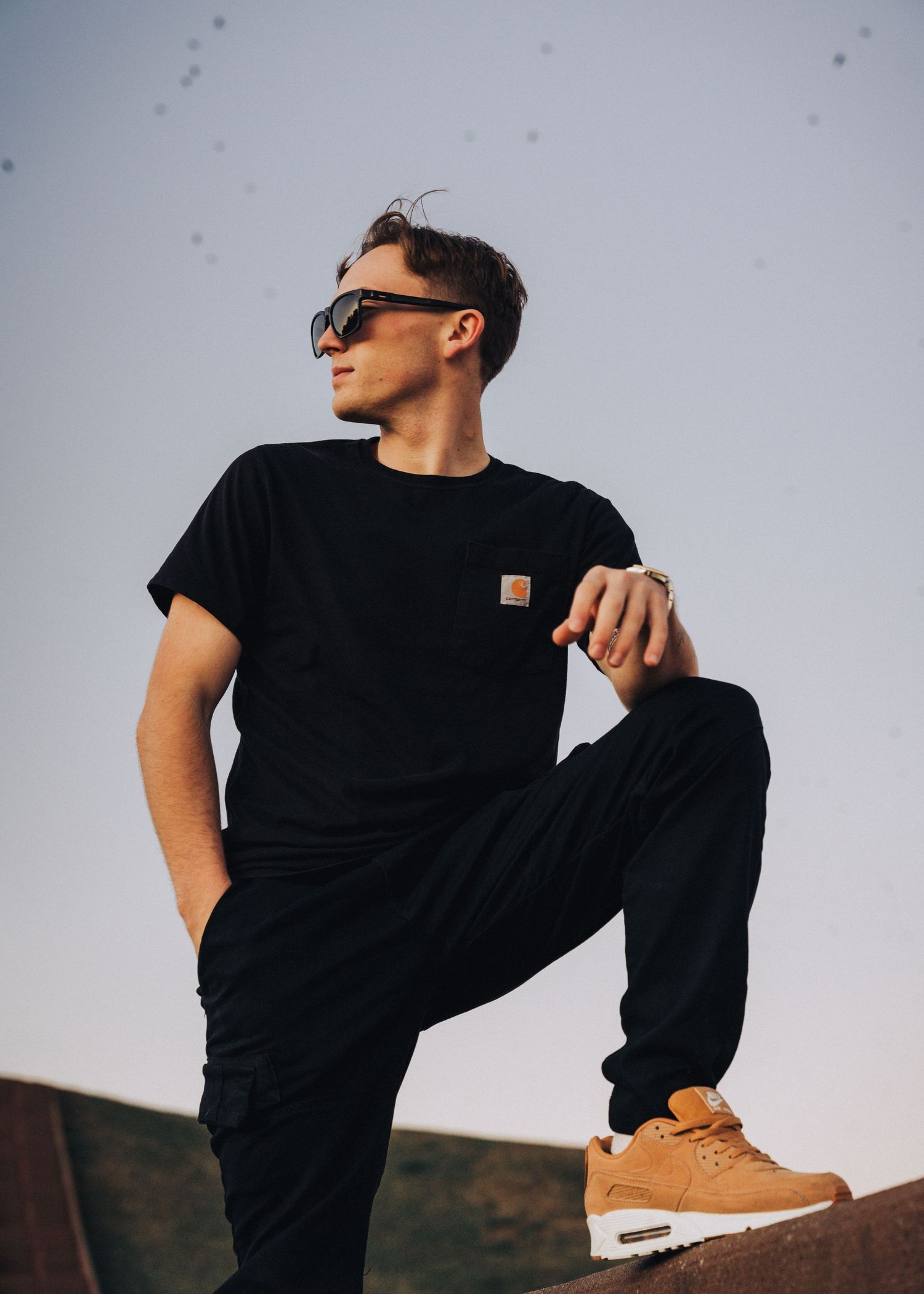 Stylish portrait of person in black outfit and tan sneakers posing on elevated surface.