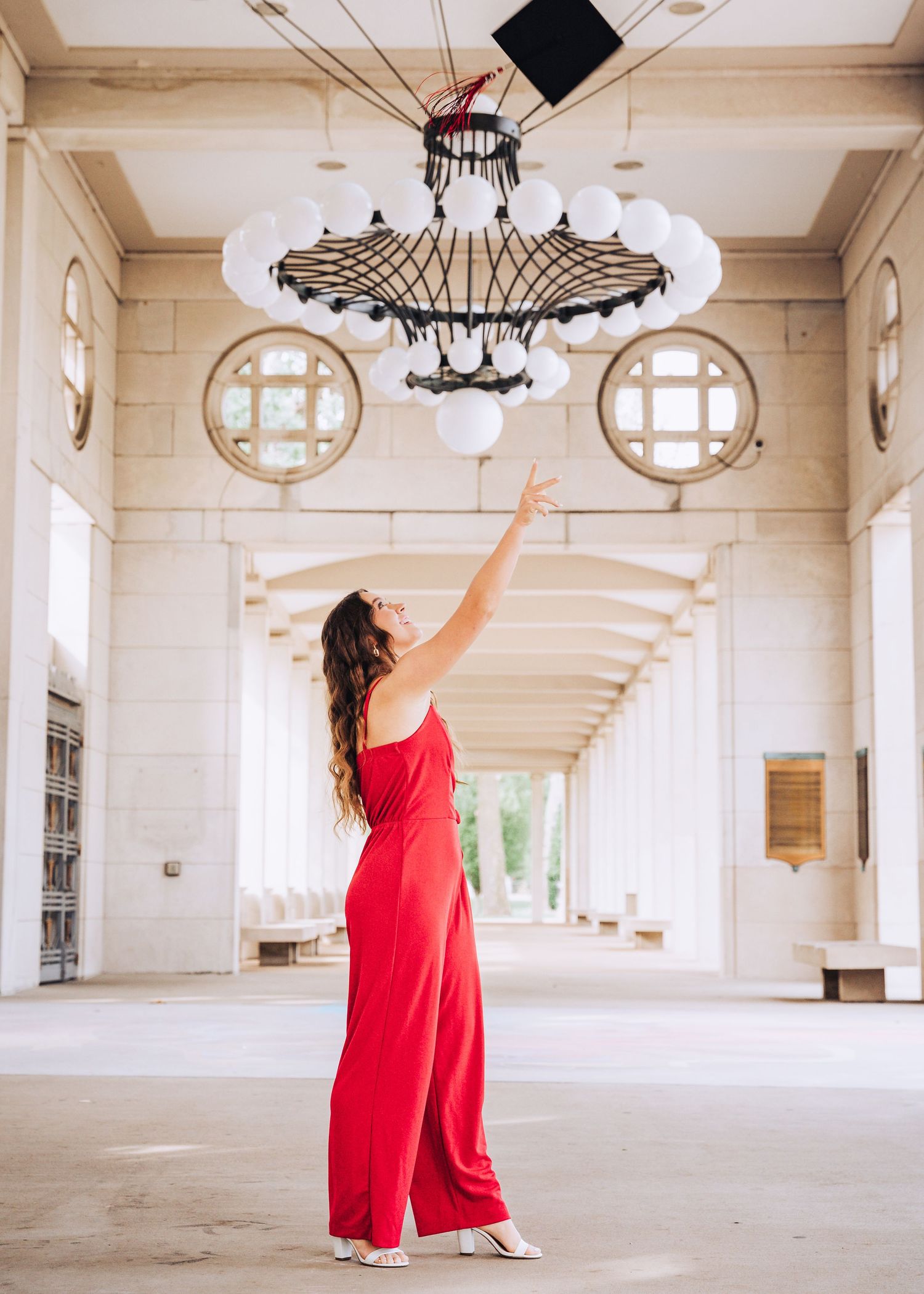 Woman in red jumpsuit reaching up toward ornate chandelier in elegant hallway.
