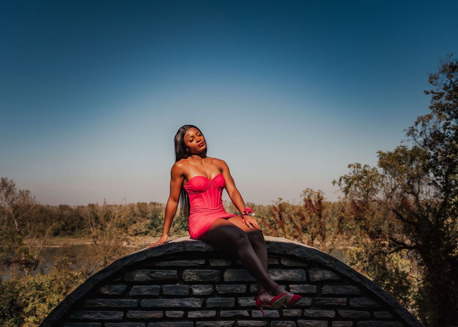 A vibrant photo of someone in a pink dress posing atop a stone wall against a blue sky.