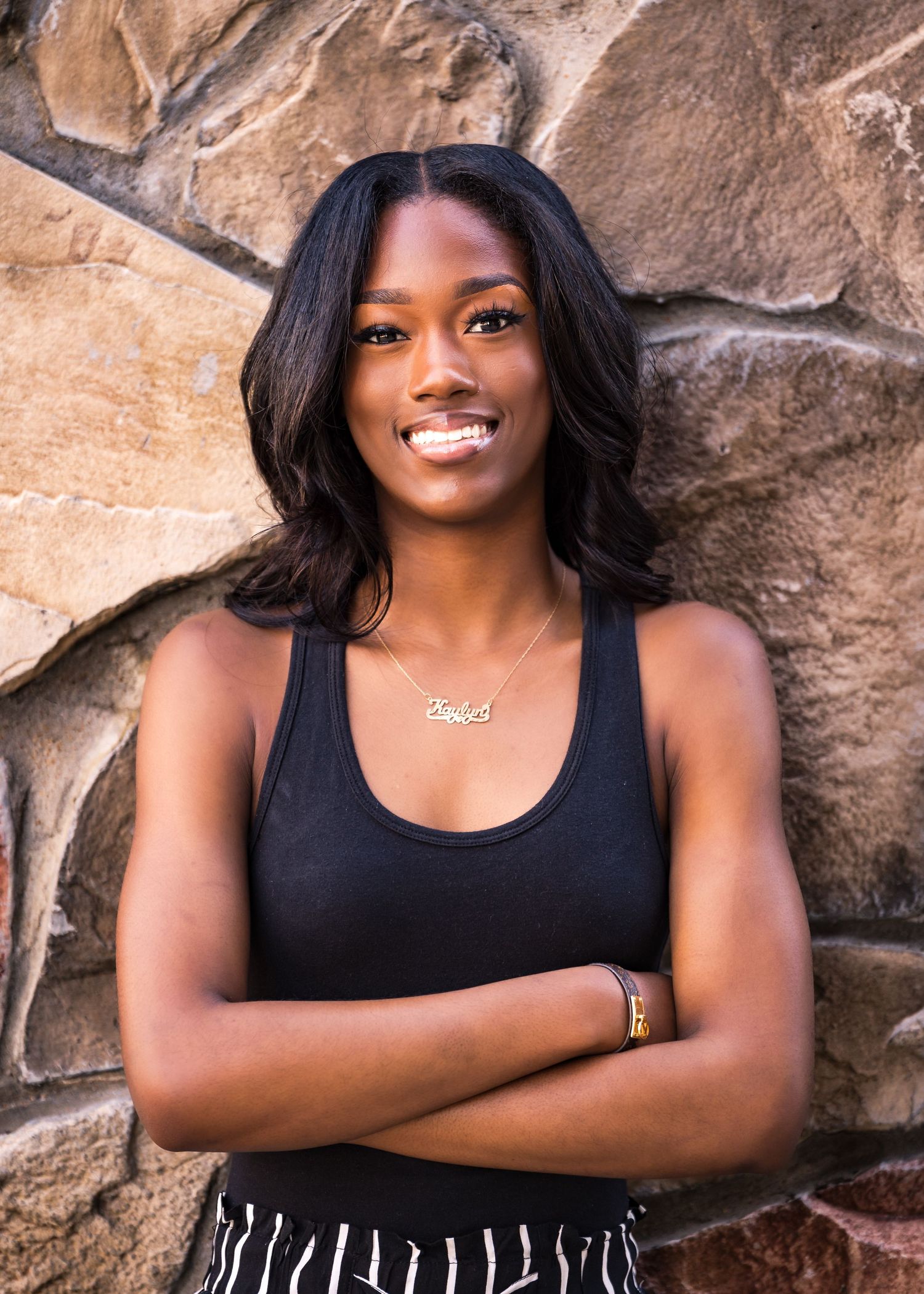 Athletic figure in black tank top poses confidently against stone wall backdrop.