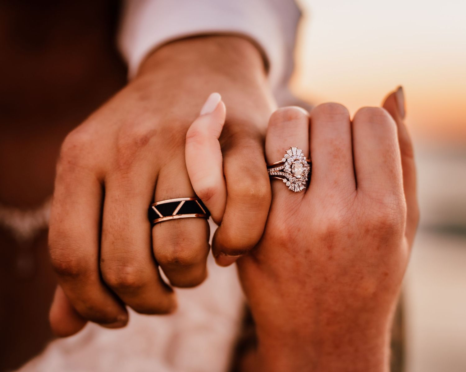 Close-up of two hands wearing wedding rings intertwined against sunset backdrop.