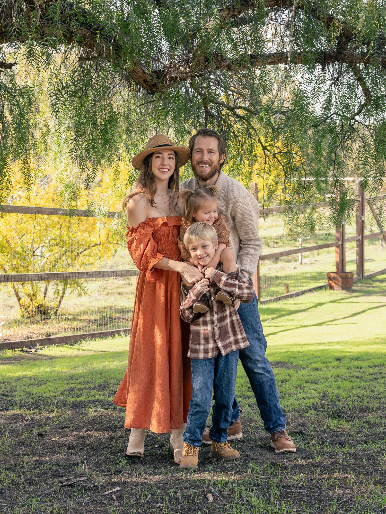 A happy family of four poses outdoors under a tree in a lush green yard during autumn.