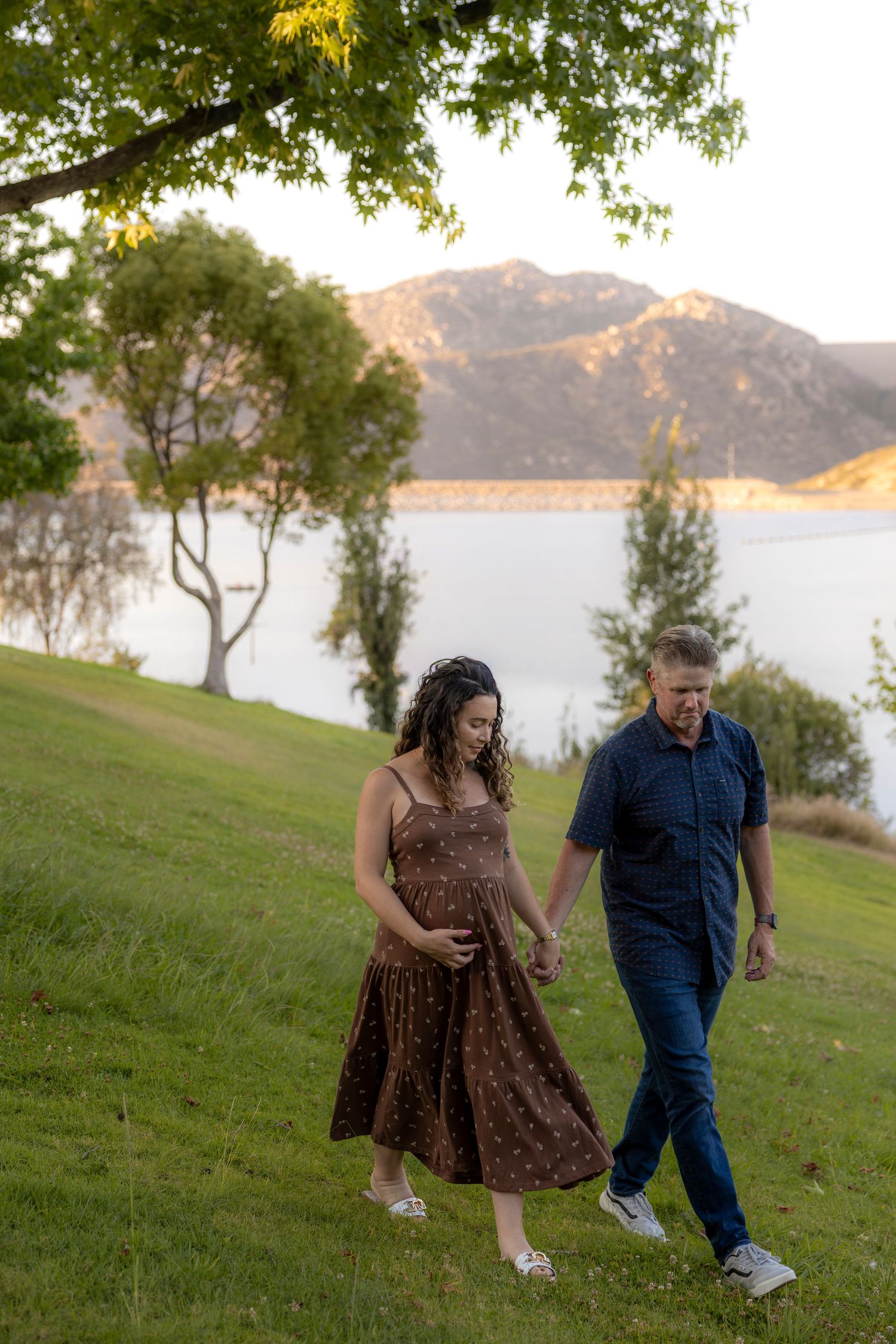 Pregnant woman and partner walking hand-in-hand on a grassy hillside with a scenic mountain lake backdrop. San Diego photographer