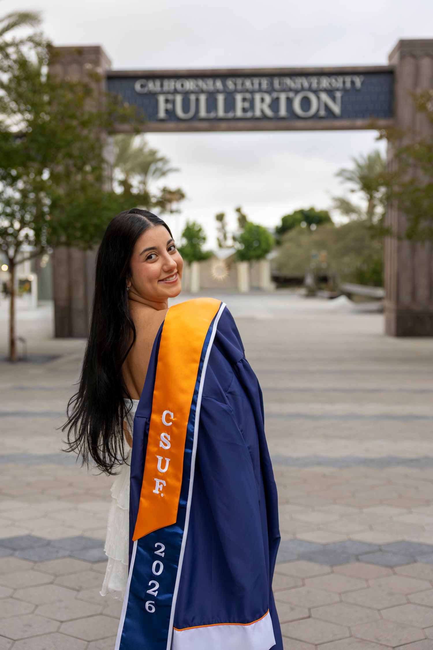 A smiling CSUF graduate poses with blue gown and orange sash in front of the California State University Fullerton sign. San Diego photograp
