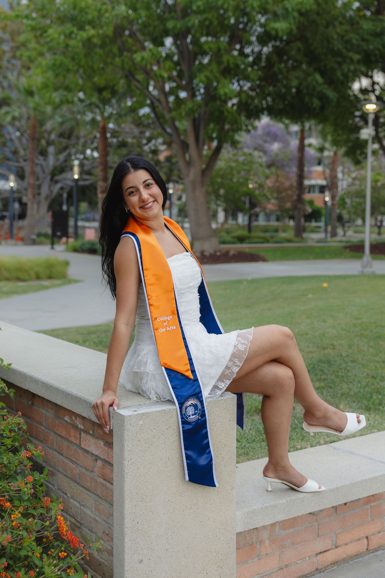Young woman in white dress with orange and blue graduation sash sits on stone wall in campus park setting. San Diego photographer