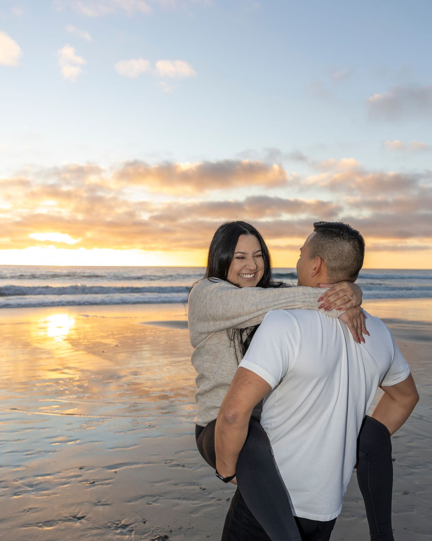 Couple embracing on a beach at sunset, woman smiling as man lifts her with ocean waves in background.