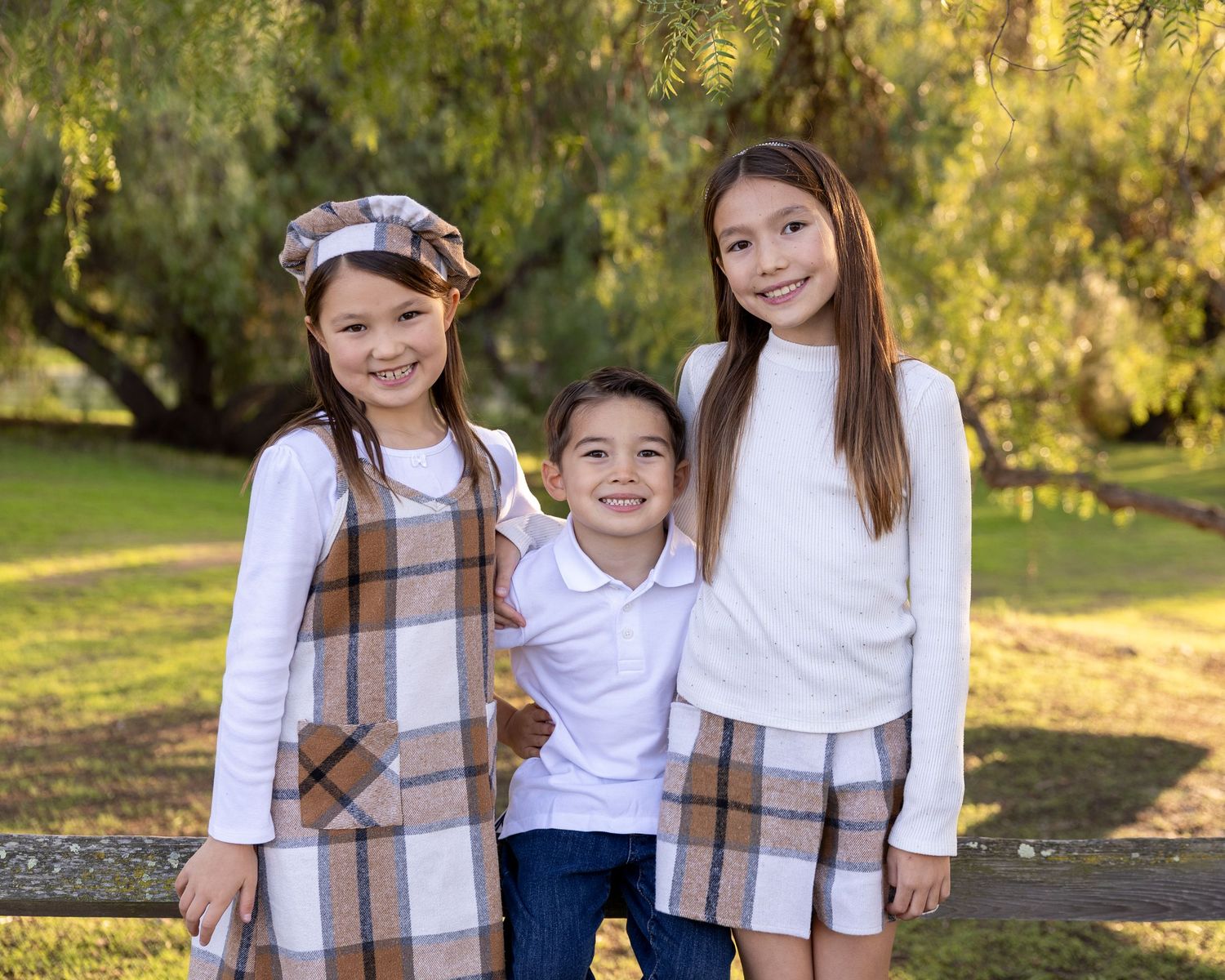 Three smiling children pose outdoors in matching plaid outfits, with lush green trees in the background.