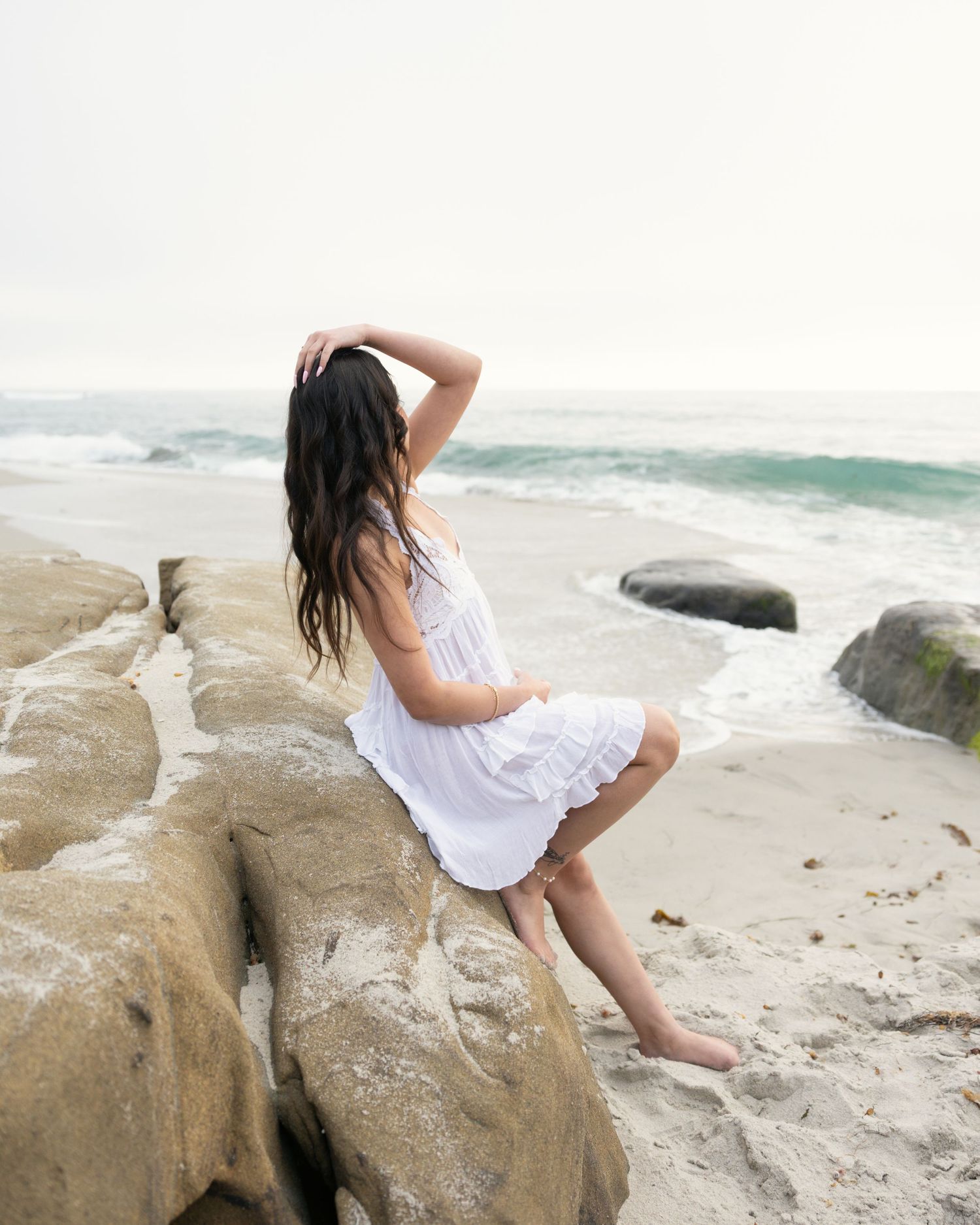 A barefoot woman in a white dress sits on coastal rocks, gazing at ocean waves on a misty beach day.