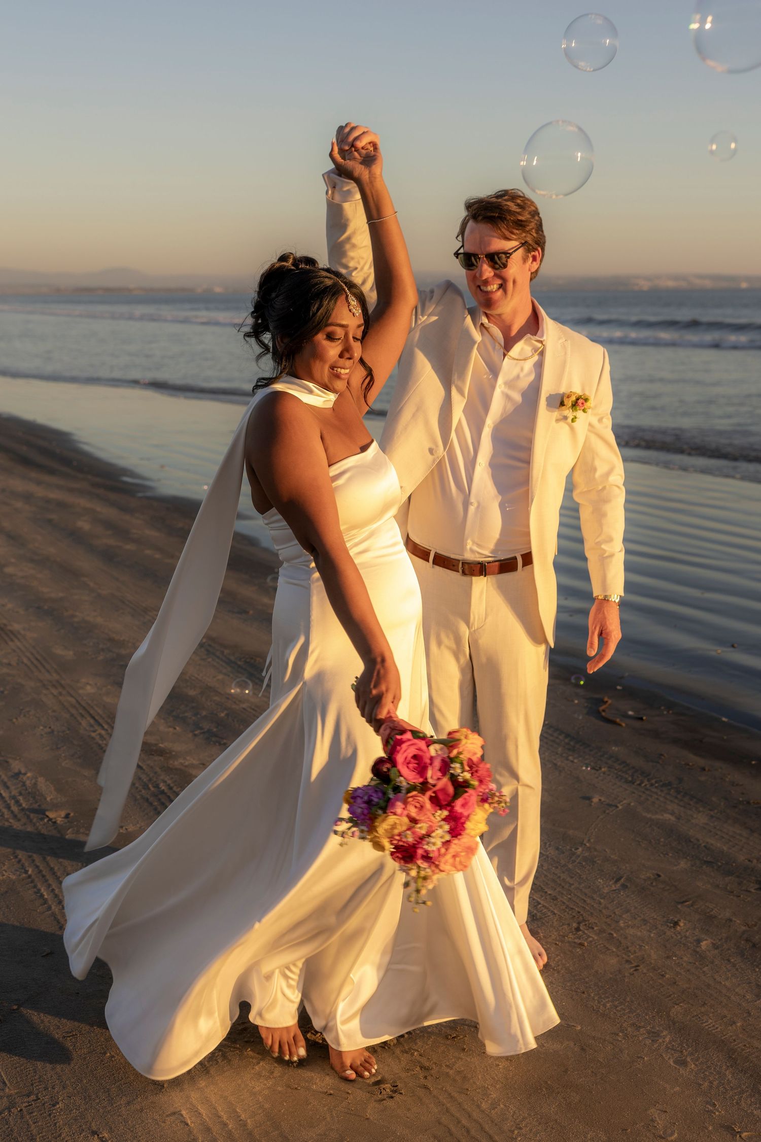 Newlywed couple celebrating on a beach at sunset, bride in white gown holding colorful bouquet, groom in white suit with sunglasses.