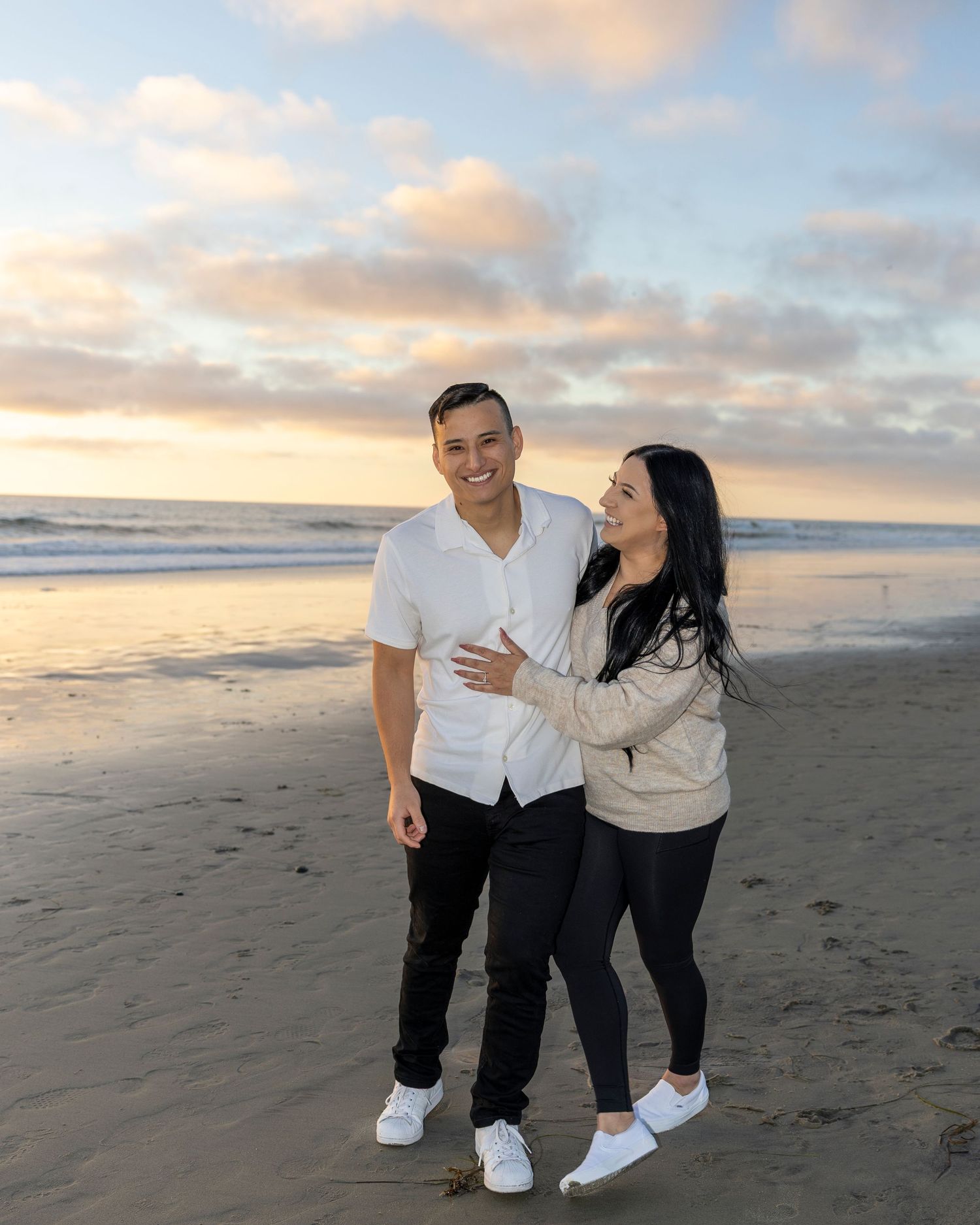 Couple smiling and embracing on a sandy beach at sunset with colorful clouds and ocean waves in the background.