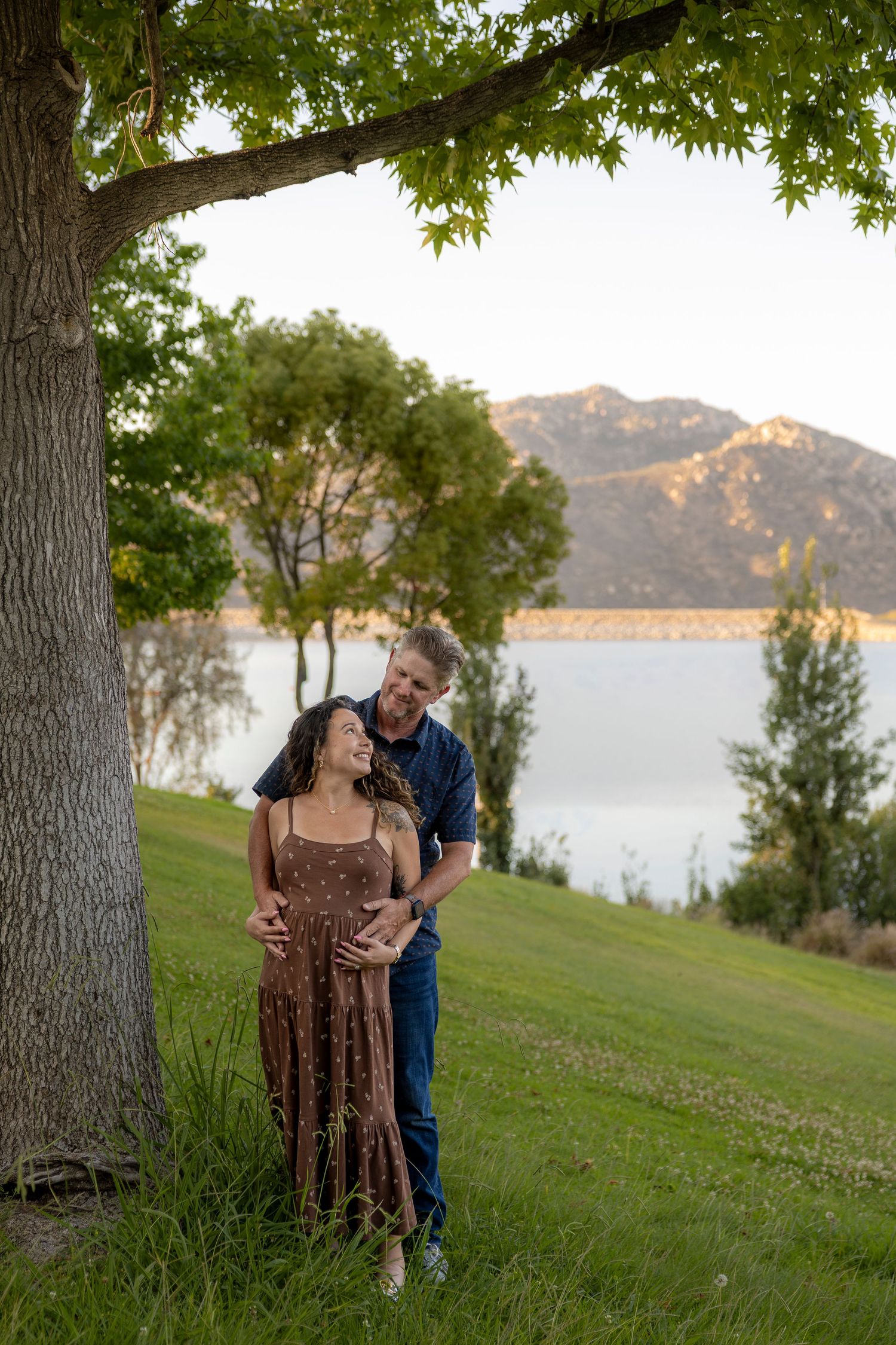 Expecting couple standing under a large tree by a mountain lake, lovingly cradling the pregnant belly. San Diego photographer